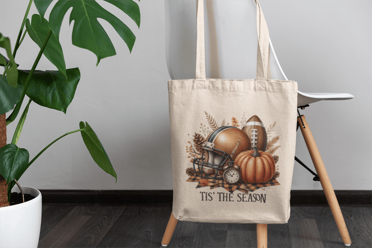 The Tis’ the Season Tote Bag by Mirabilia Boutique, a beige canvas market bag with fall football and pumpkin design, hangs on a white chair near a green potted plant.