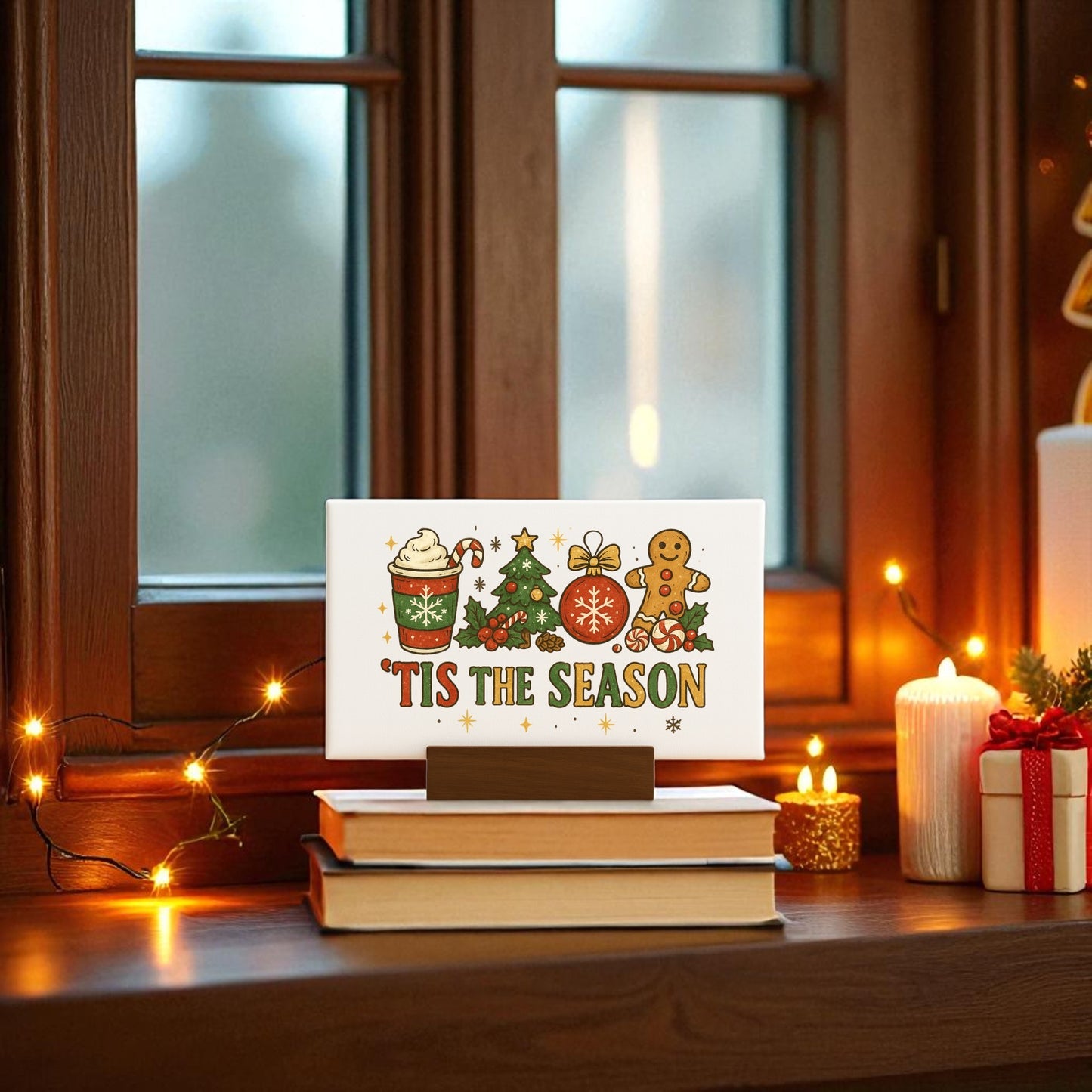 The 'Tis the Season Mini Canvas | 6x4 Christmas Sign with Base by Mirabilia Boutique sits on stacked books near a window, surrounded by fairy lights, candles, and festive holiday décor.