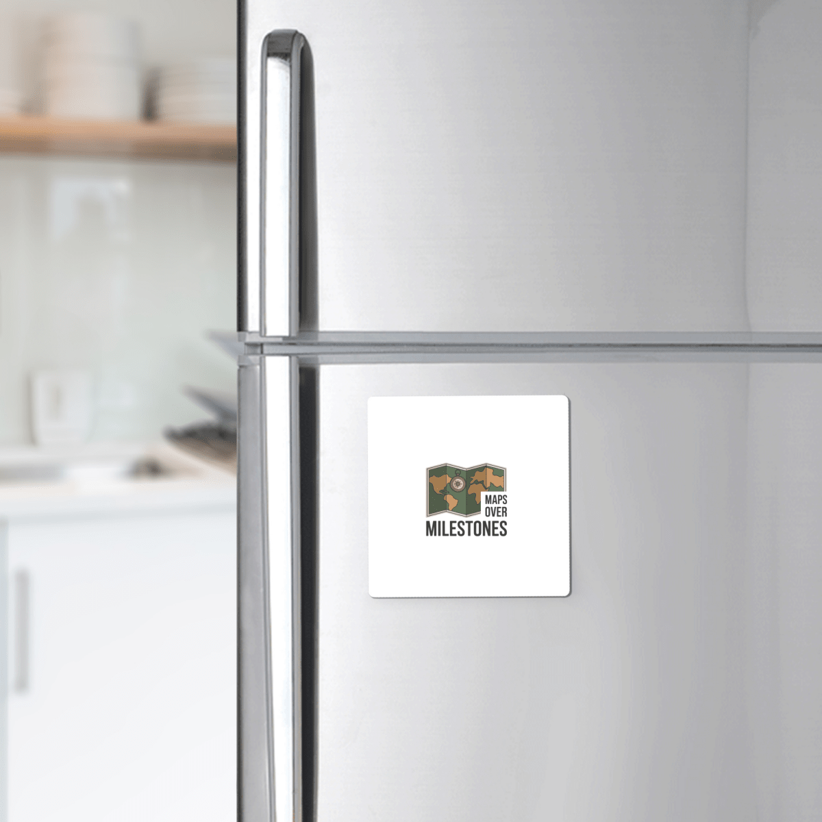 The Maps Over Milestones Travel Magnet by Mirabilia Boutique, featuring a logo with a map and the phrase "MAPS OVER MILESTONES," adds adventure-inspired style to any stainless steel fridge. Blurred kitchen shelves and dishes appear in the background.