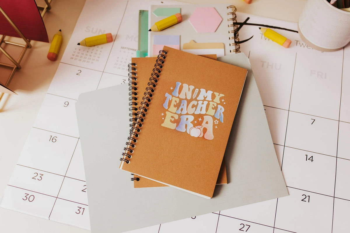 The Mirabilia Boutique "In My Teacher Era" Aesthetic Teacher Spiral Journal is a retro notebook, perfect as a Back to School teacher gift, pictured with other notebooks, highlighters, and a large desk calendar.
