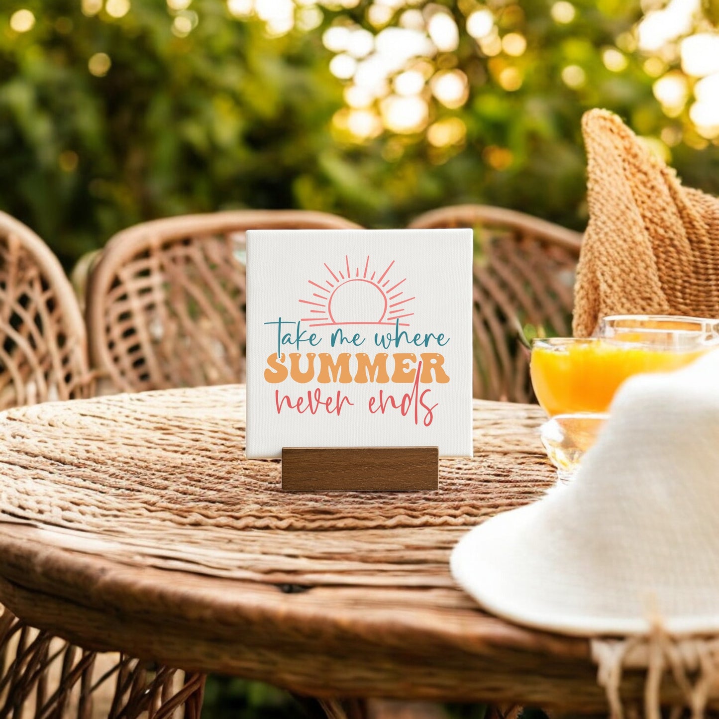 The Mirabilia Boutique "Take Me Where Summer Never Ends" Mini Canvas sits on a wicker table beside orange juice and a white hat, perfect for beach house decor with lush greenery softly blurred in the background.