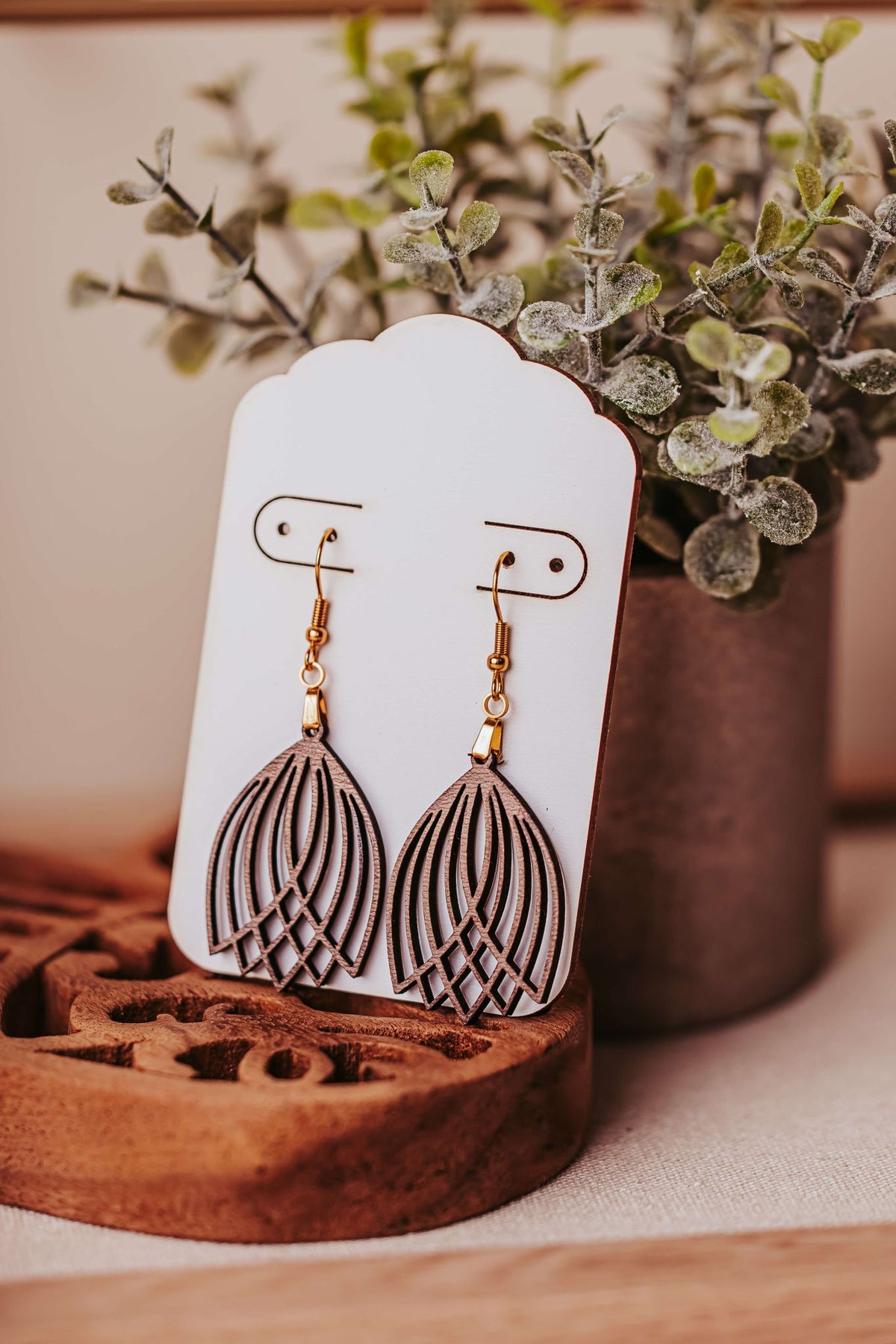 The Mirabilia Boutique Laser Cut Tulip Earrings are lightweight, gold and grey boho dangle wooden statement jewelry, shown layered on a white card with a potted plant and wooden tray in the background.