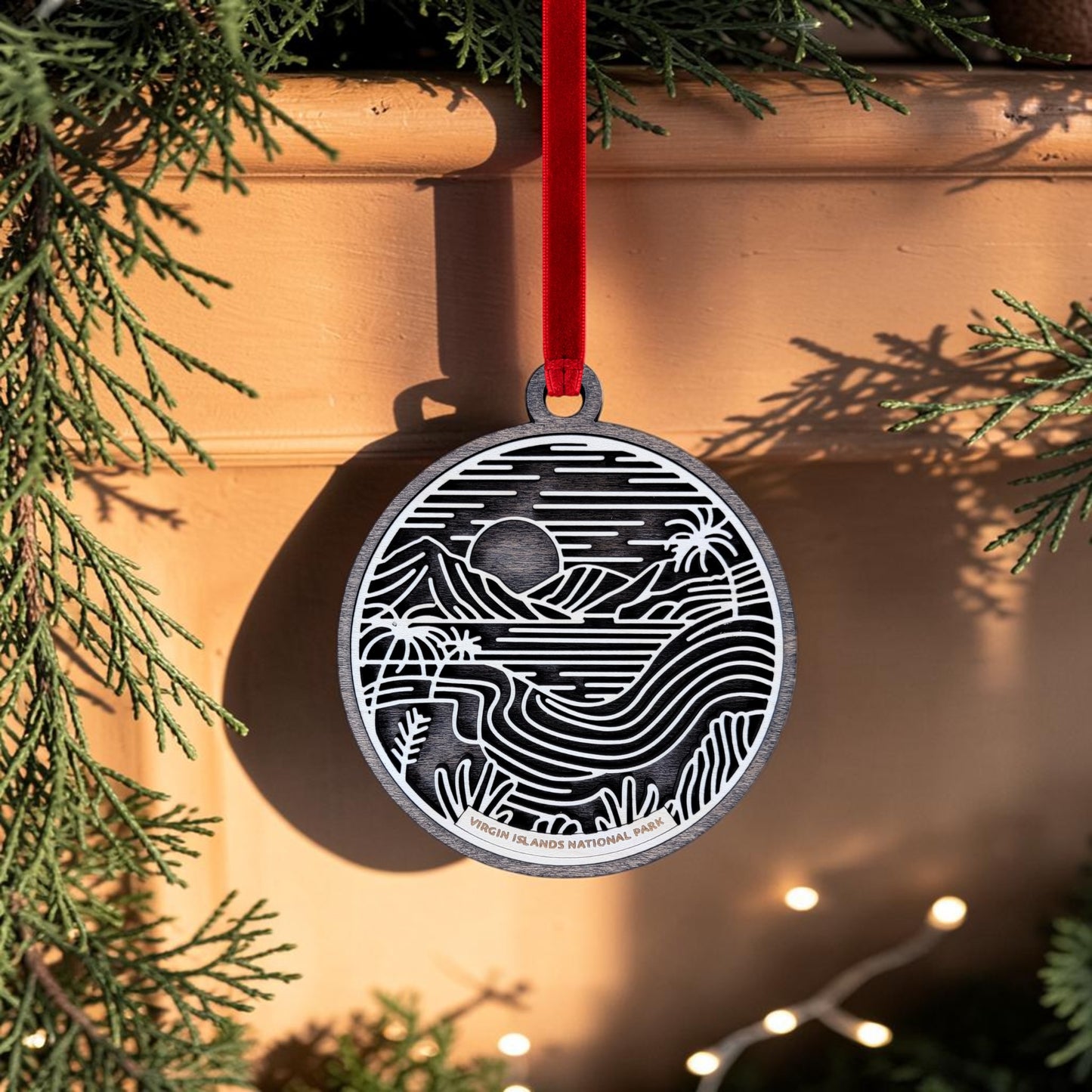 The Virgin Islands National Park Line Art Christmas Ornament by Mirabilia Boutique, featuring a scenic landscape design, hangs from a red ribbon amid evergreen branches, with sunlight casting shadows and a few twinkling lights below.