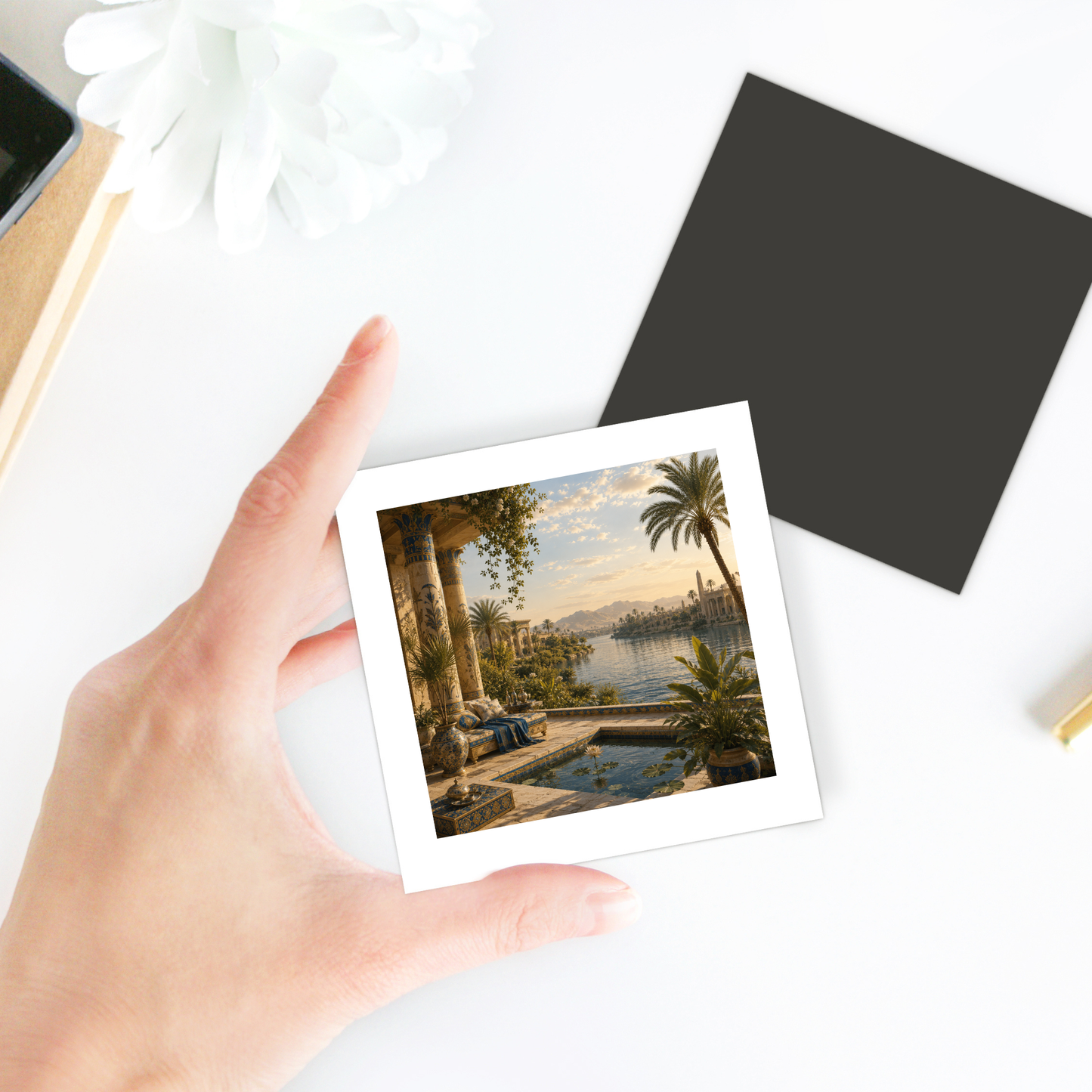 A hand holds the “View of the Nile Magnet | Ancient Egyptian Art” by Mirabilia Boutique, featuring an ancient riverside scene with palm trees and columns; a black square and a white flower rest on the white table nearby.