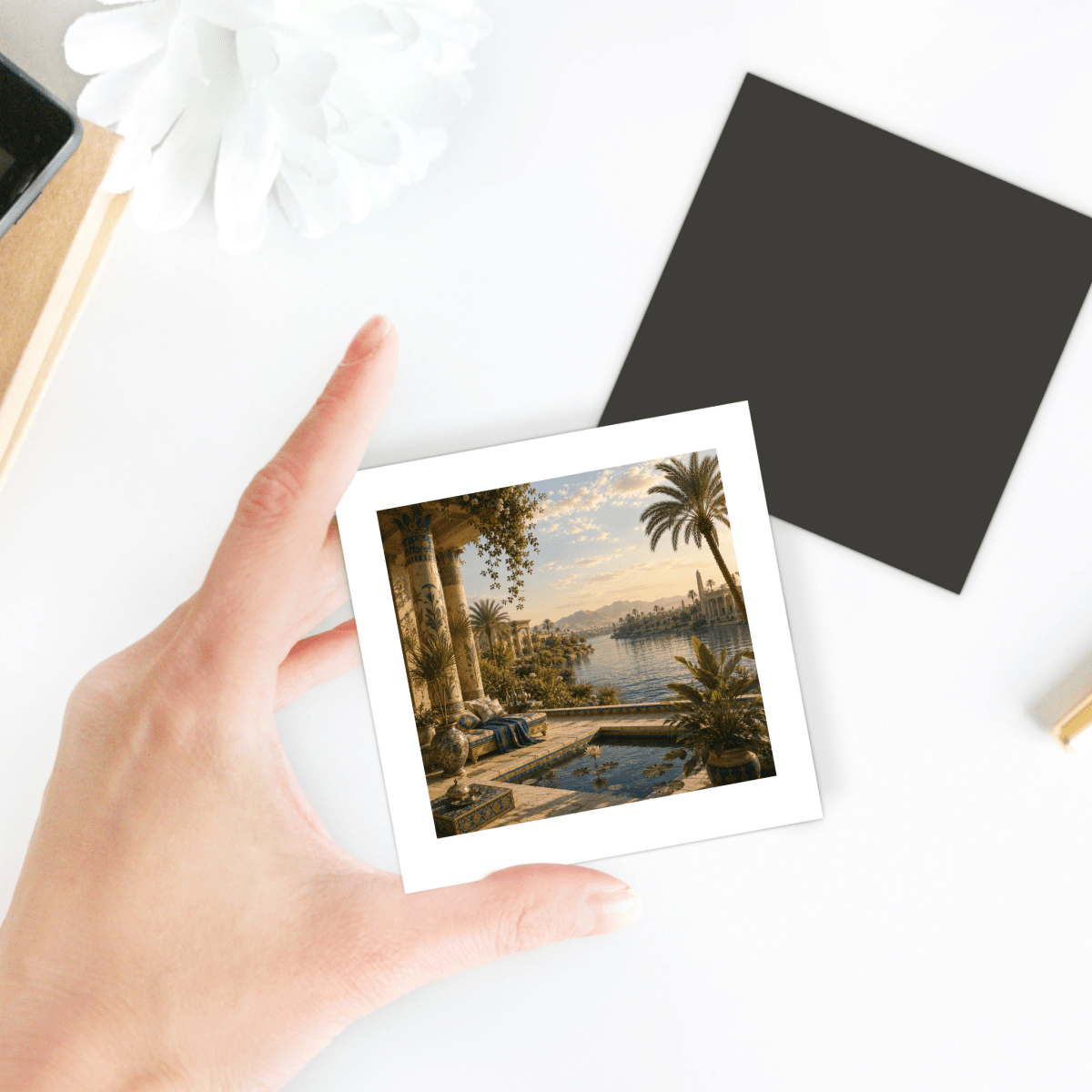 A hand holds the “View of the Nile Magnet | Ancient Egyptian Art” by Mirabilia Boutique, featuring an ancient riverside scene with palm trees and columns; a black square and a white flower rest on the white table nearby.