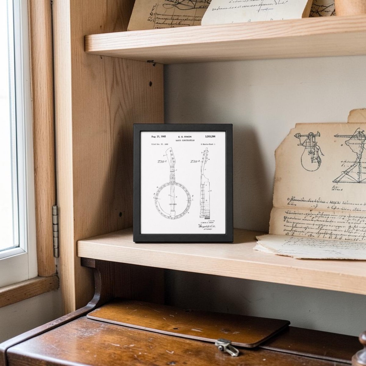 The Banjo Patent Print 8x10 Wall Art by Mirabilia Boutique is displayed on a bookshelf near a window, alongside old documents and sketches, with a wooden desk positioned below.