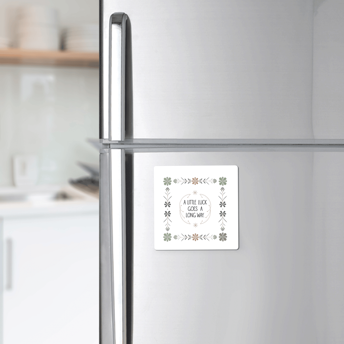 A Mirabilia Boutique "A Little Luck Goes a Long Way" magnet with a green floral border decorates the fridge door, while shelves with dishes appear in the background.