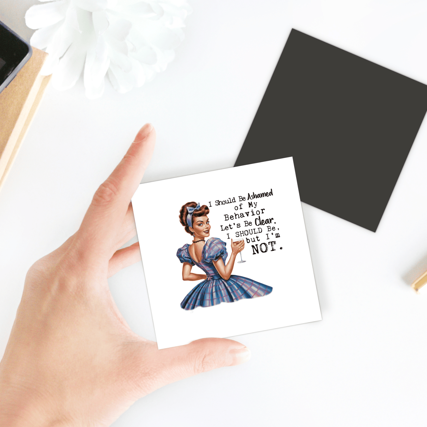 A hand holds the Mirabilia Boutique “I Should Be Ashamed” 3x3 UV printed magnet with a sassy retro woman and funny text—a bold, cheeky gift for wine lovers or party girls. Black square card and white flower are also shown nearby.