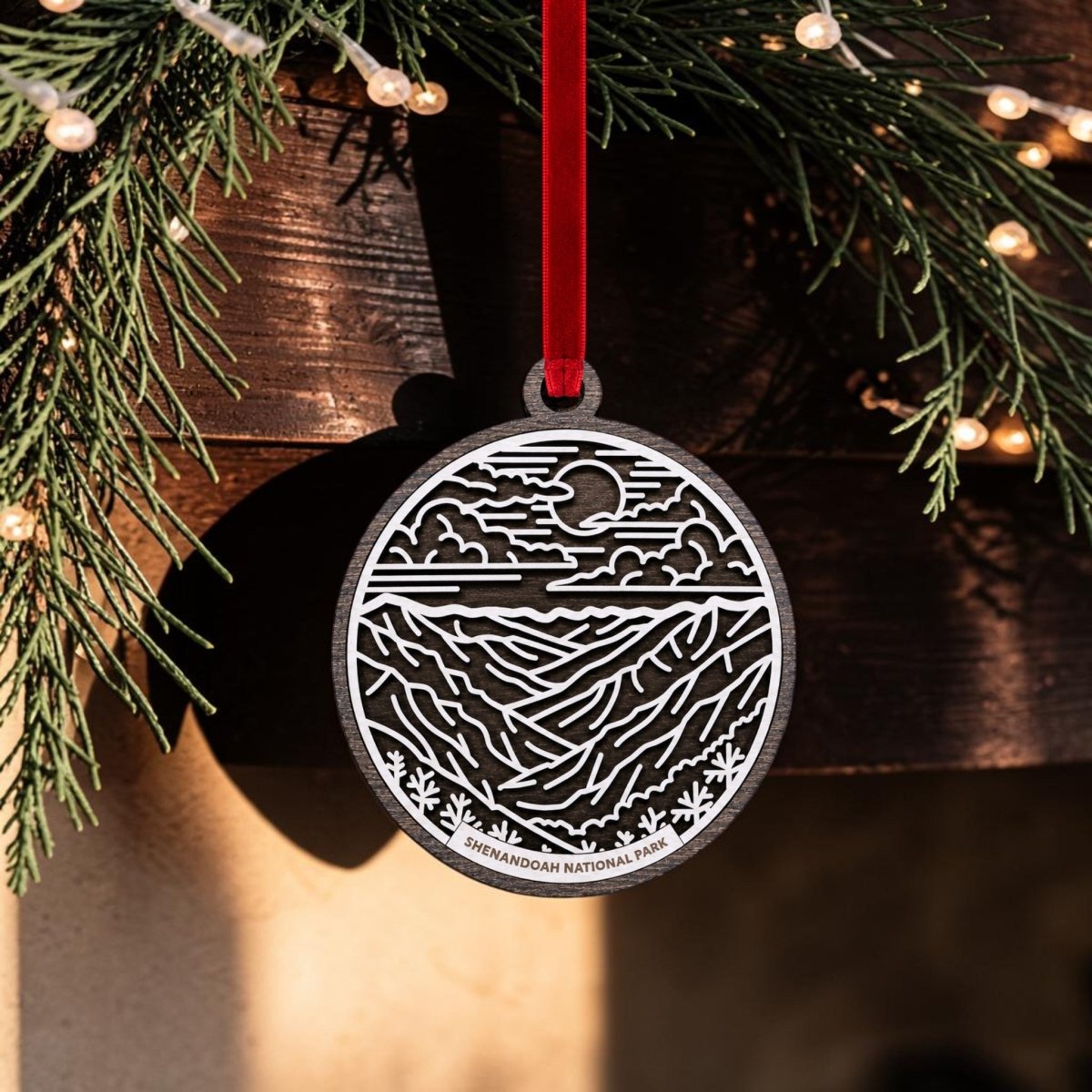 A round metal Shenandoah National Park Line Art Christmas Ornament by Mirabilia Boutique hangs from a red ribbon, showcasing a mountain and sky design. The background features evergreen branches and string lights, while the ornament displays "SHENANDOAH NATIONAL PARK" at the bottom, inspired by Blue Ridge Mountains art.