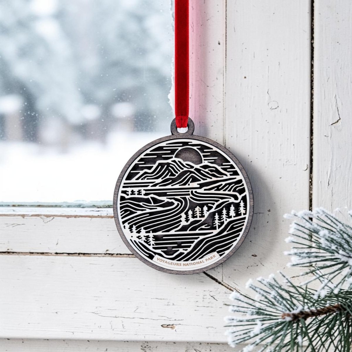 The Mirabilia Boutique Voyageurs National Park Line Art Christmas Ornament, a handcrafted wooden oval featuring a black and white landscape design, hangs from a red ribbon on a white window frame; pine branches and snow are visible outside.