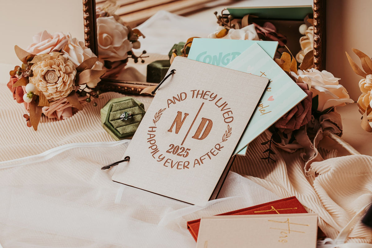 The Wedding Card Keepsake Binder | Personalized Memory Book by Mirabilia Boutique, featuring "And They Lived Happily Ever After N/D 2025" on the cover, is displayed on fabric with flowers, a green ring box, cards, and a decorative mirror.