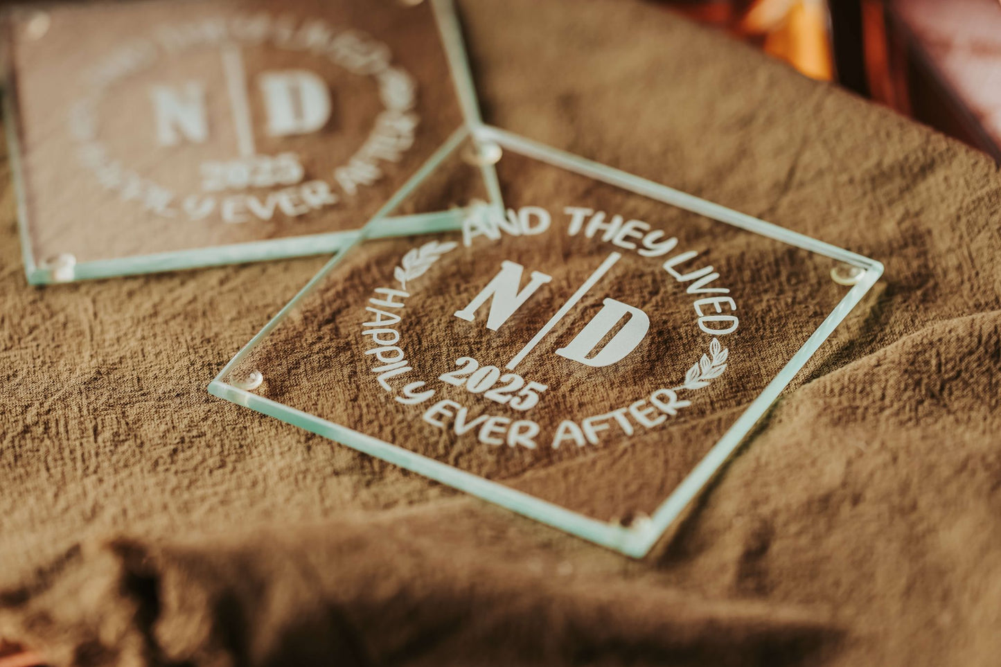A Personalized Wedding Coaster Favor by Mirabilia Boutique, engraved with "N | D 2025 AND THEY LIVED HAPPILY EVER AFTER," rests on brown textured fabric, while another custom-made glass coaster is softly blurred in the background.