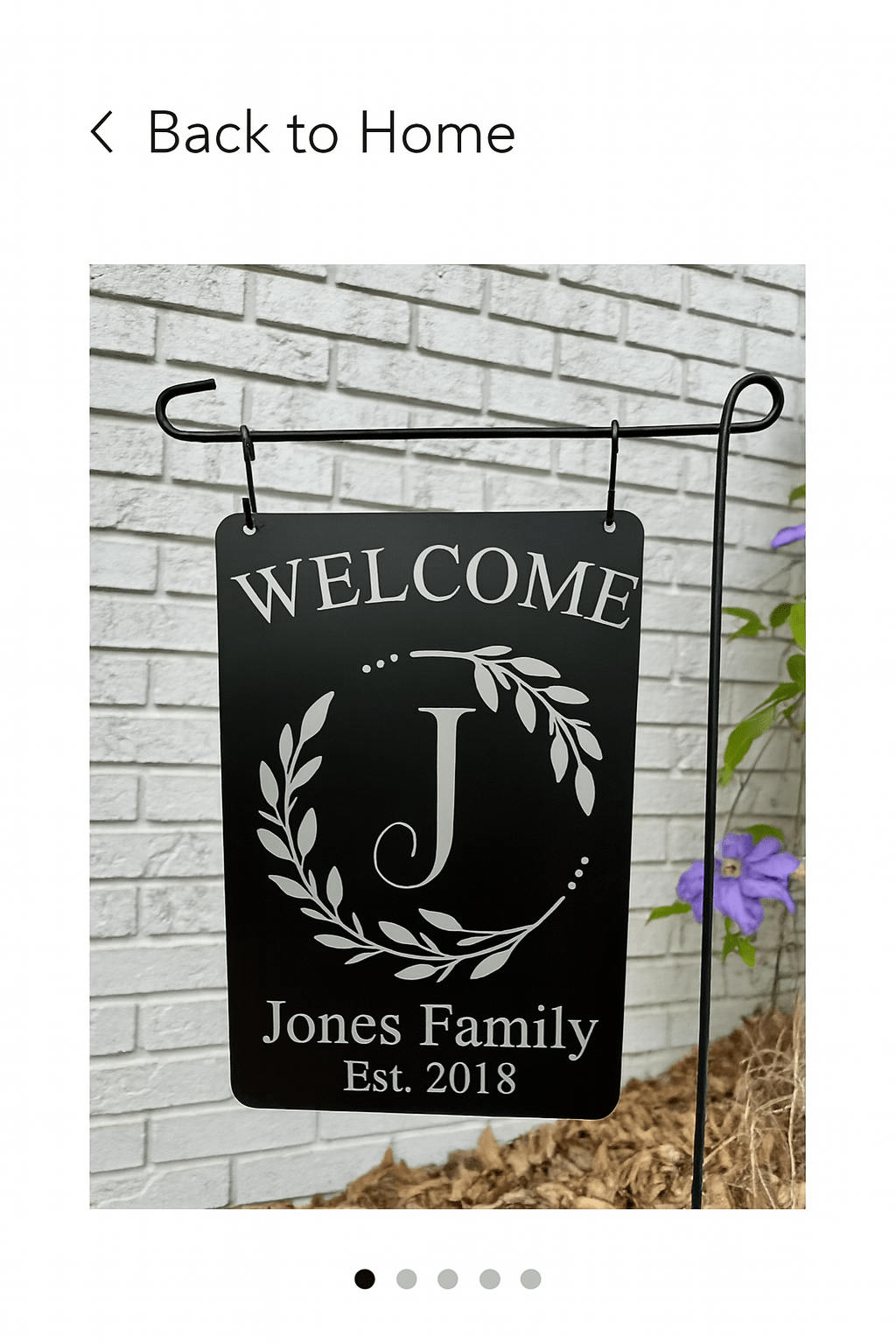 The Mirabilia Boutique Personalized Welcome Garden Sign features "Welcome," a large "J," laurel branches, and "Jones Family Est. 2018." This custom family yard decor makes a thoughtful housewarming gift and looks lovely by flowers or a white brick wall.