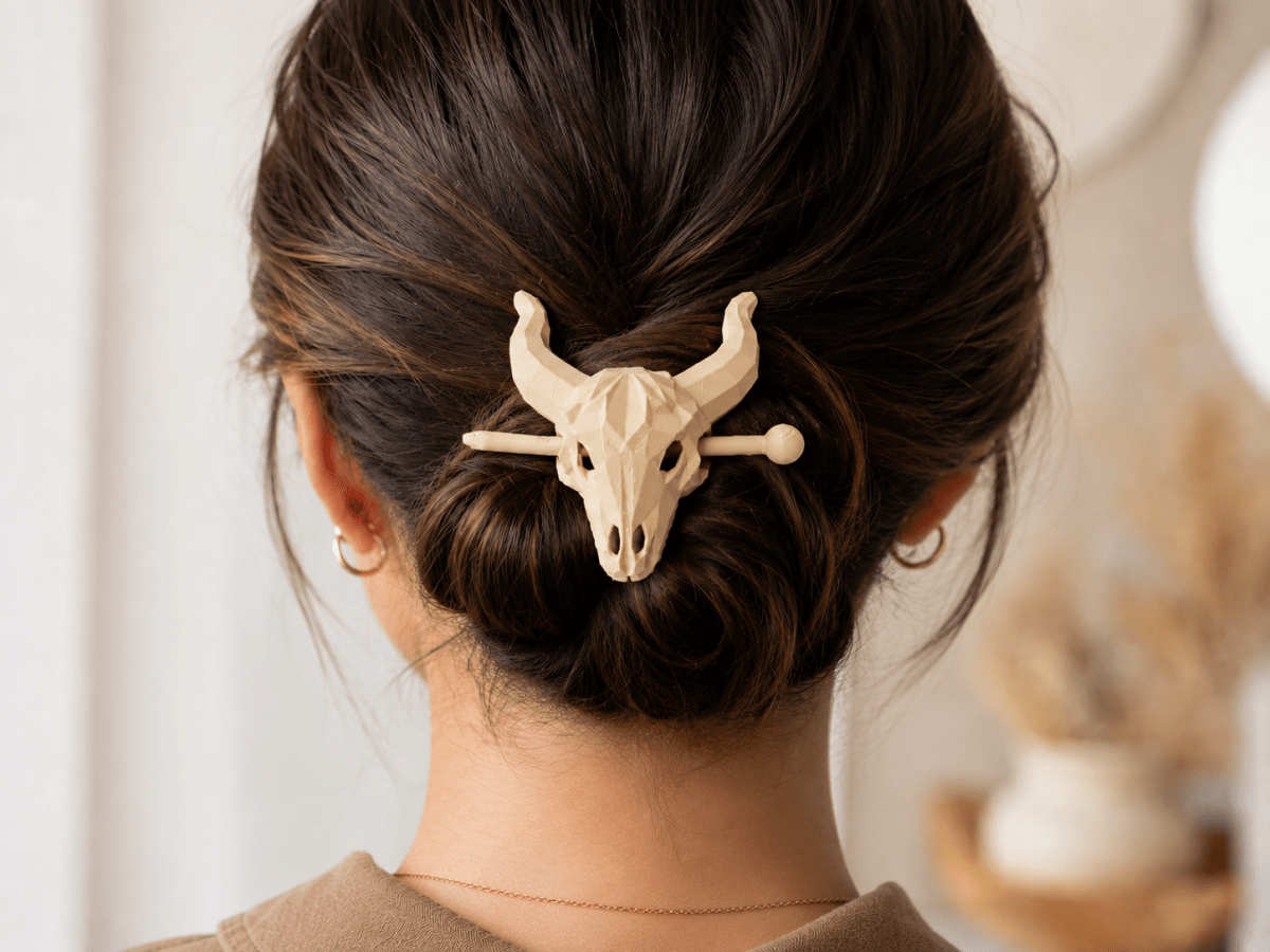 A woman’s dark hair is styled in a bun and held with the Mirabilia Boutique Longhorn Skull Hairpin | Western Theme Hair Accessory in beige. She pairs it with small gold hoop earrings and a brown top, against a softly blurred background.
