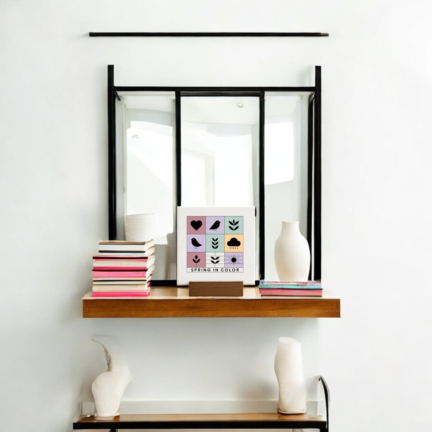 A modern wall shelf displays books, two white vases, and the Mirabilia Boutique "Spring in Color" Mini Canvas Sign with Wood Base, featuring colorful spring icons. Above the shelf hangs a black-trimmed mirror.