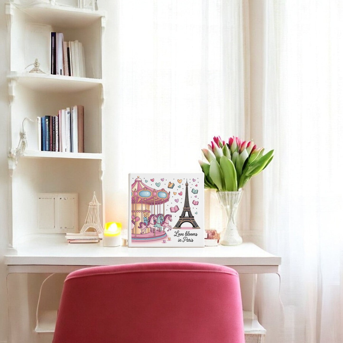 A white desk with a pink chair sits by a window with sheer curtains. On the desk are tulips, a candle, and Mirabilia Boutique’s “Love Blooms in Paris Canvas – Eiffel Tower & Carousel Art,” perfect for Paris art fans.