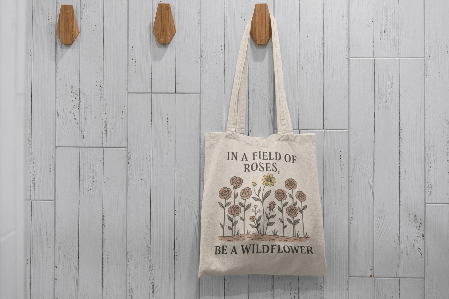 The Be a Wildflower Tote Bag | Inspirational Canvas Tote by Mirabilia Boutique hangs on a hook against a gray wall, featuring flower illustrations and "In a field of roses, be a wildflower" on eco-friendly cotton canvas.