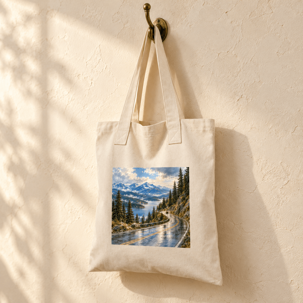 The Scenic Byway Tote Bag hanging on a hook, featuring a print of a winding mountain road alongside a lake.