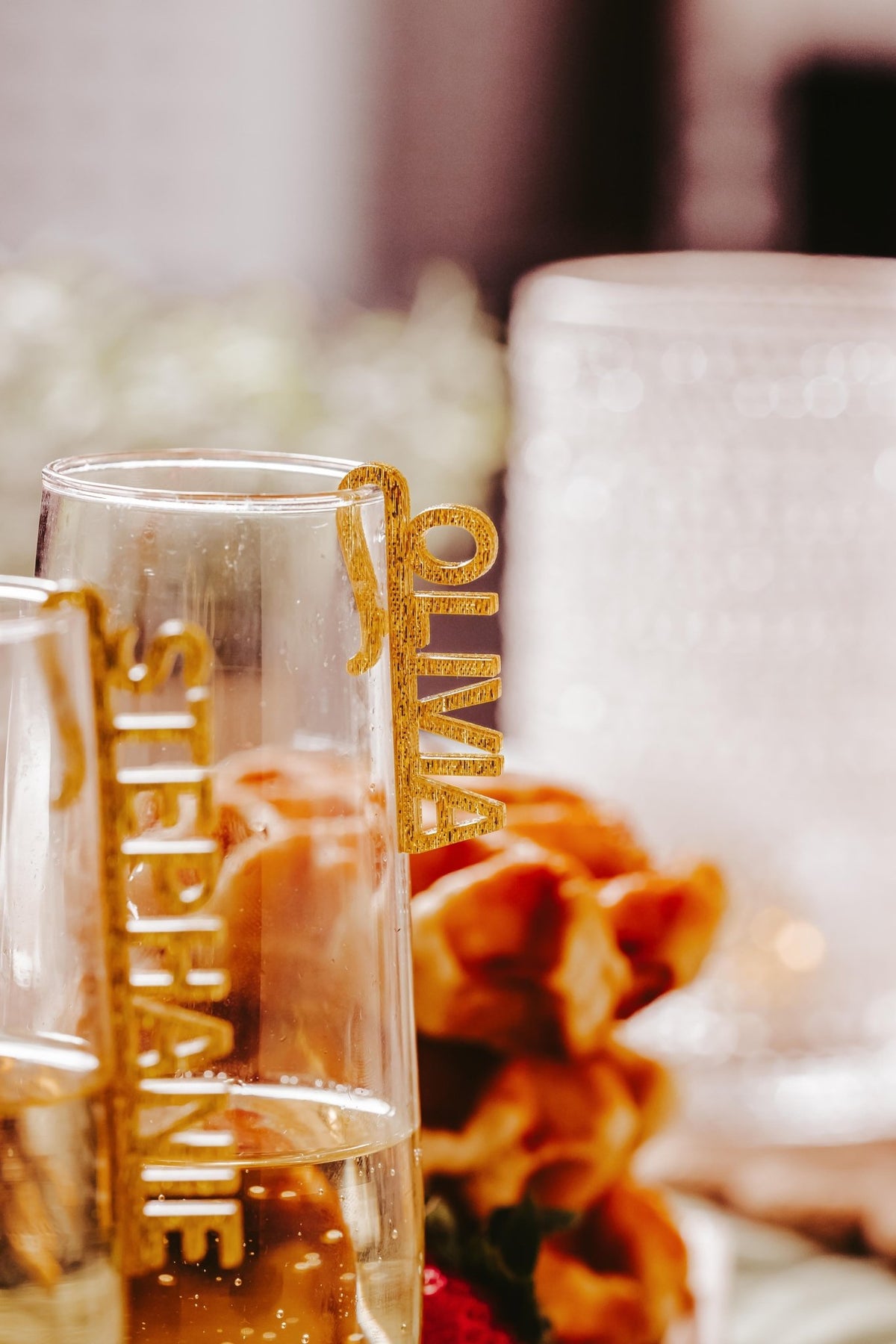 Two champagne glasses feature gold custom acrylic wine glass name charms by Mirabilia Boutique—one reading "STEPHANIE," the other "OLIVIA." Personalized drink tags add elegance, perfect for weddings, bachelorette parties, or bridal shower décor.