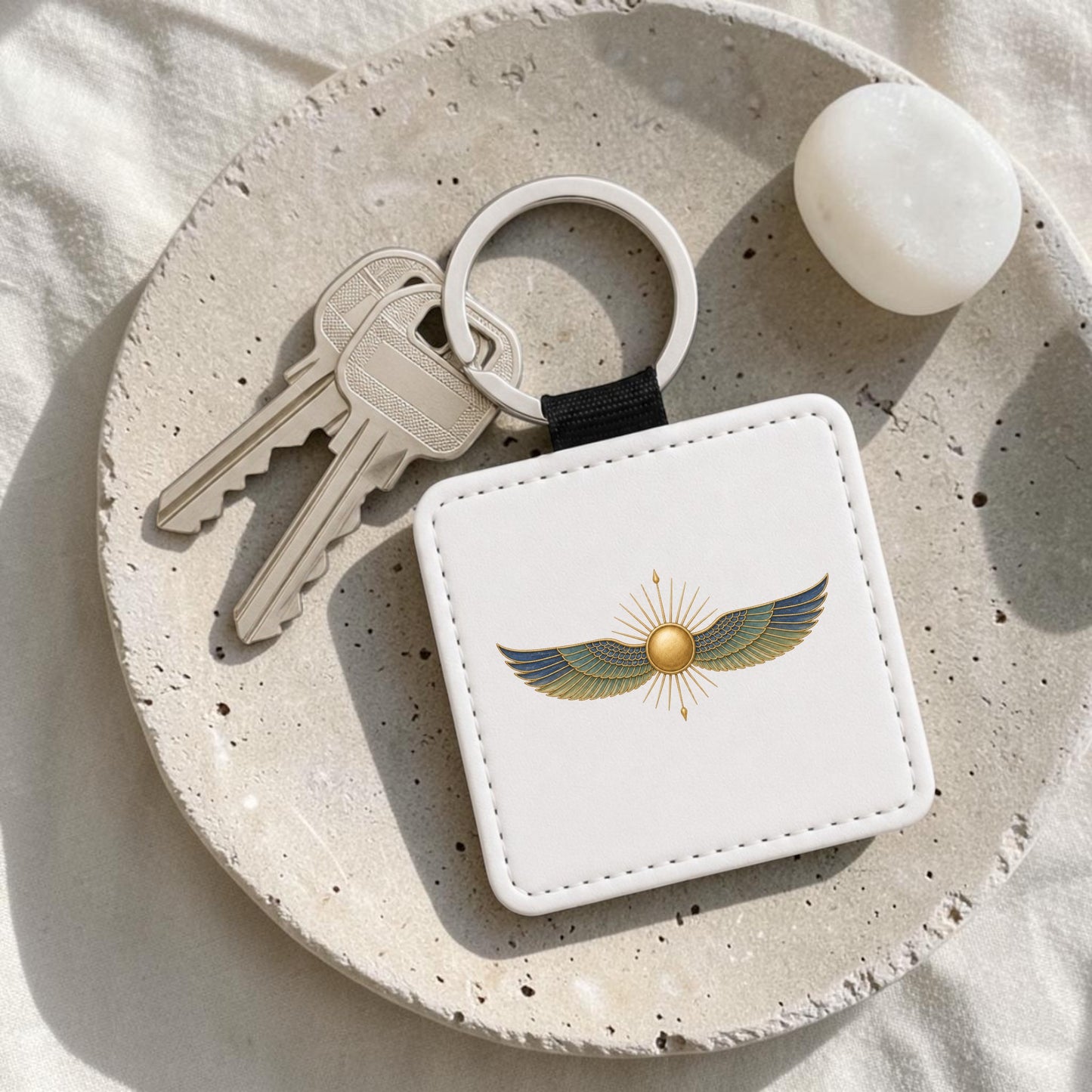 A Winged Sun Keychain | Egyptian Inspired Keepsake by Mirabilia Boutique, featuring a white square holder decorated with a golden sun and blue and gold wings, rests with two keys on a speckled beige plate as part of the Lapis & Lotus Collection. A smooth white stone is also placed on the plate.