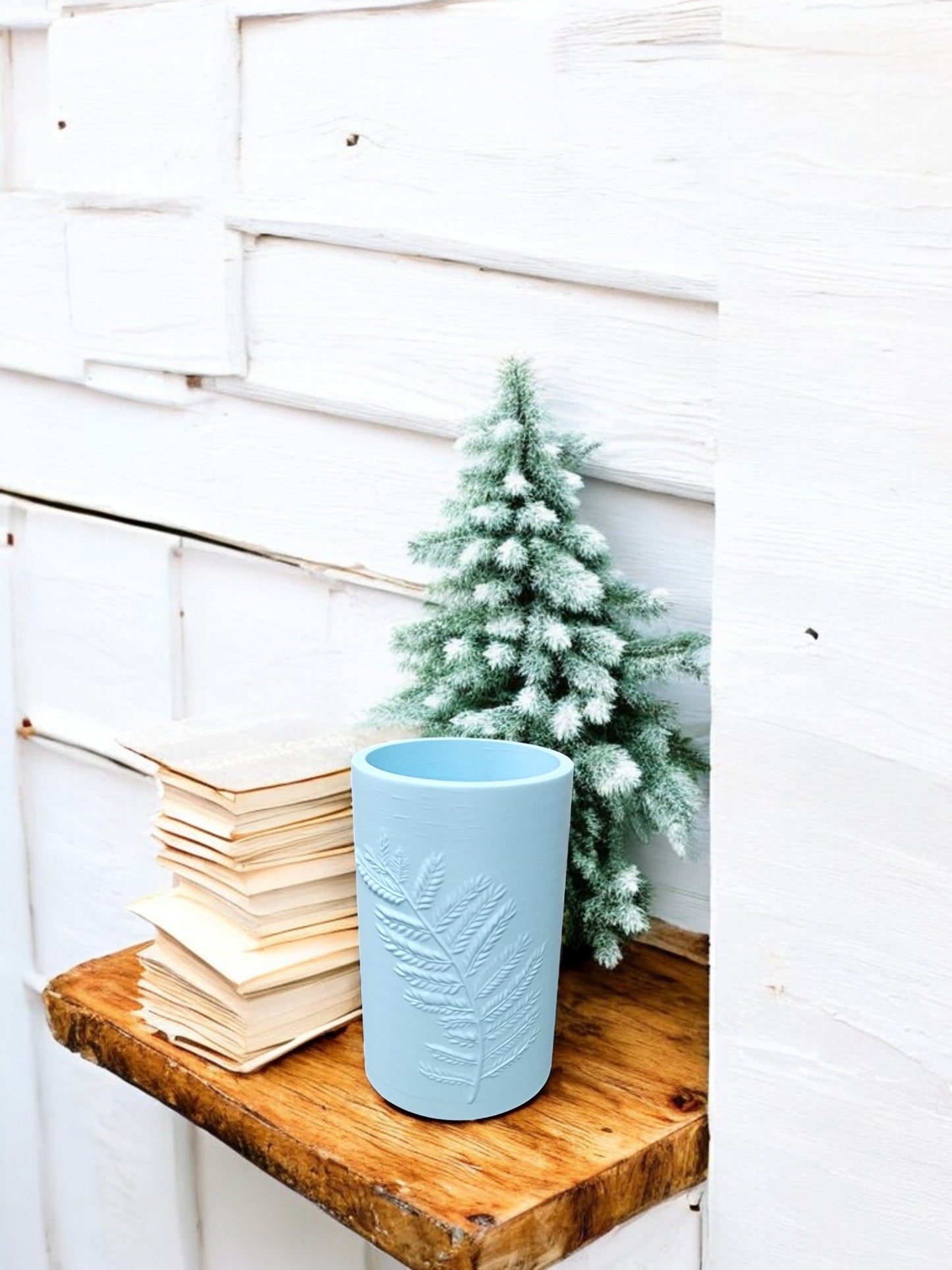 A small frosted pine tree, Mirabilia Boutique’s Glacial Blue Winter Fern Vase – Woodland Accent, and a stack of books sit on a rustic wooden shelf against a white paneled wall.