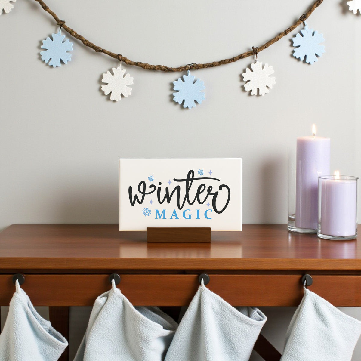 A Mirabilia Boutique Winter Magic Sign (6x4 Minimalist Winter Decor) sits on a wooden table beside two lit lavender candles. Blue and white snowflake garland hangs above, with light blue towels on hooks below.