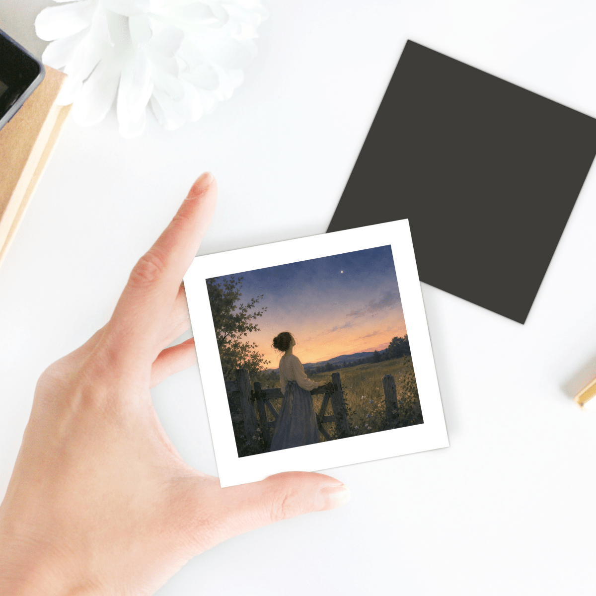 A hand holds the Mirabilia Boutique “Wonder, Always Twilight Landscape Magnet | Nature Art,” featuring a woman in a wildflower meadow at sunset, gazing at the sky. On the table are a white flower, a black square, and a notebook.