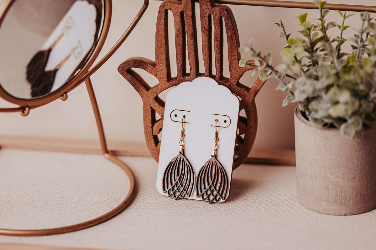 Laser Cut Tulip Earrings by Mirabilia Boutique—lightweight, modern wooden boho dangles with hypoallergenic hooks—are displayed on a white card atop a hand-shaped stand, accented by a round mirror and potted plant.