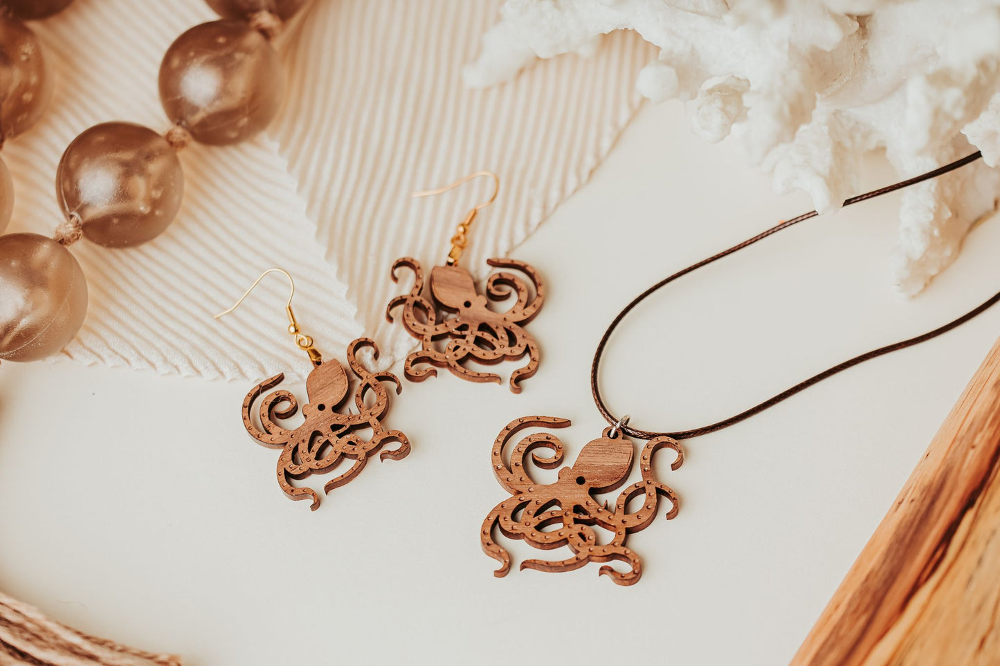 The Octopus Earrings from Mirabilia Boutique—laser-cut wood ocean jewelry perfect for sea life lovers—are shown on a light surface near large brown beads, a white coral ornament, and a matching octopus pendant necklace.