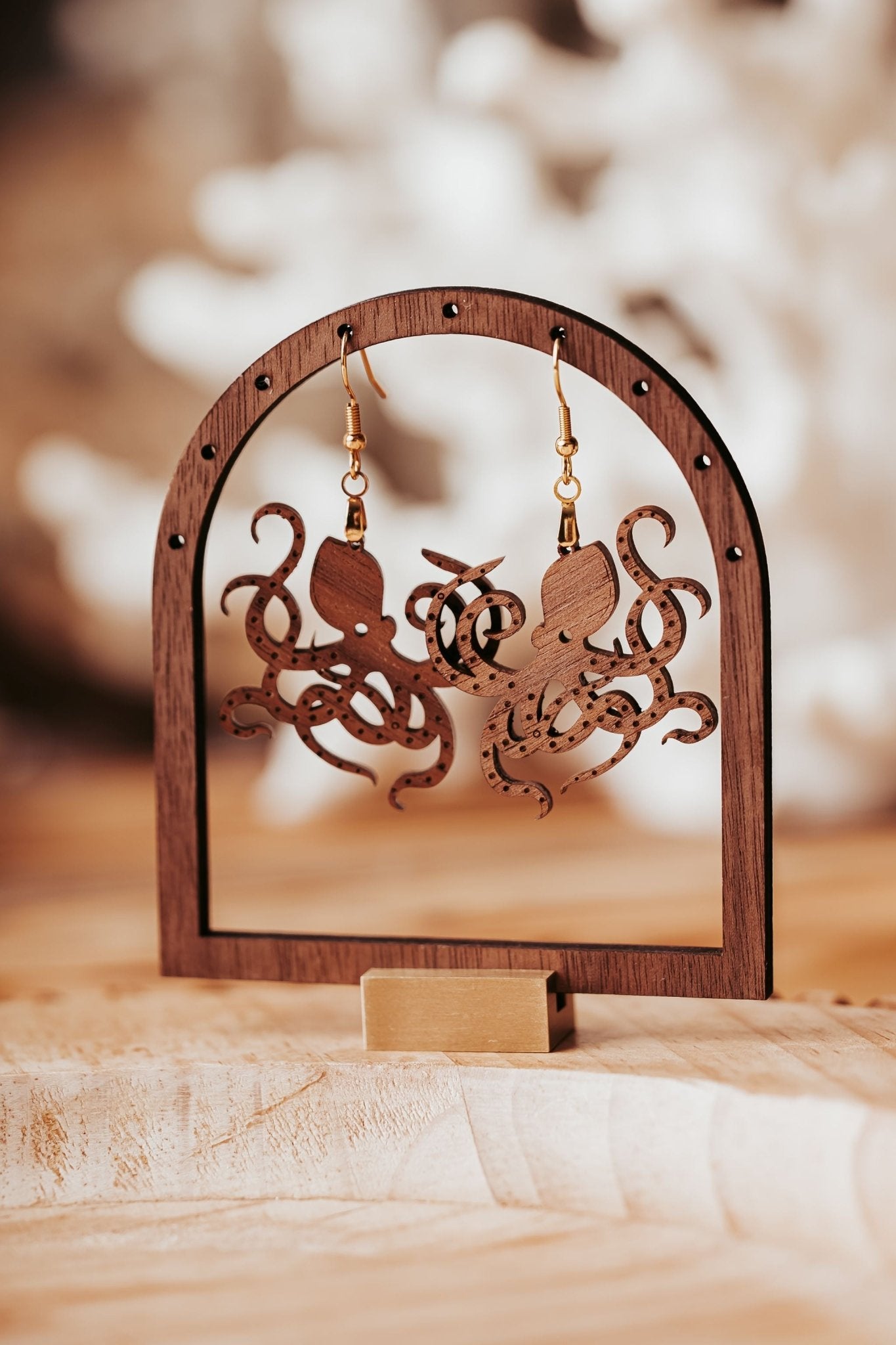 A pair of Mirabilia Boutique’s Octopus Earrings – laser cut wood ocean jewelry for sea life lovers – hang from a wooden arch display on a light surface with a blurred background.