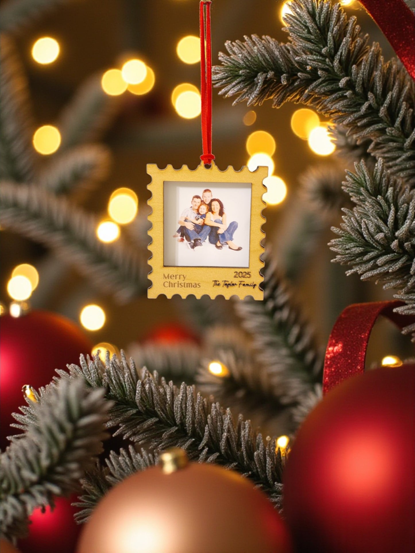 A close-up of a Christmas tree branch displays the Mirabilia Boutique Personalized Christmas Stamp Frame Ornament, featuring a custom family photo and "Merry Christmas 2023," surrounded by festive baubles and warm lights.