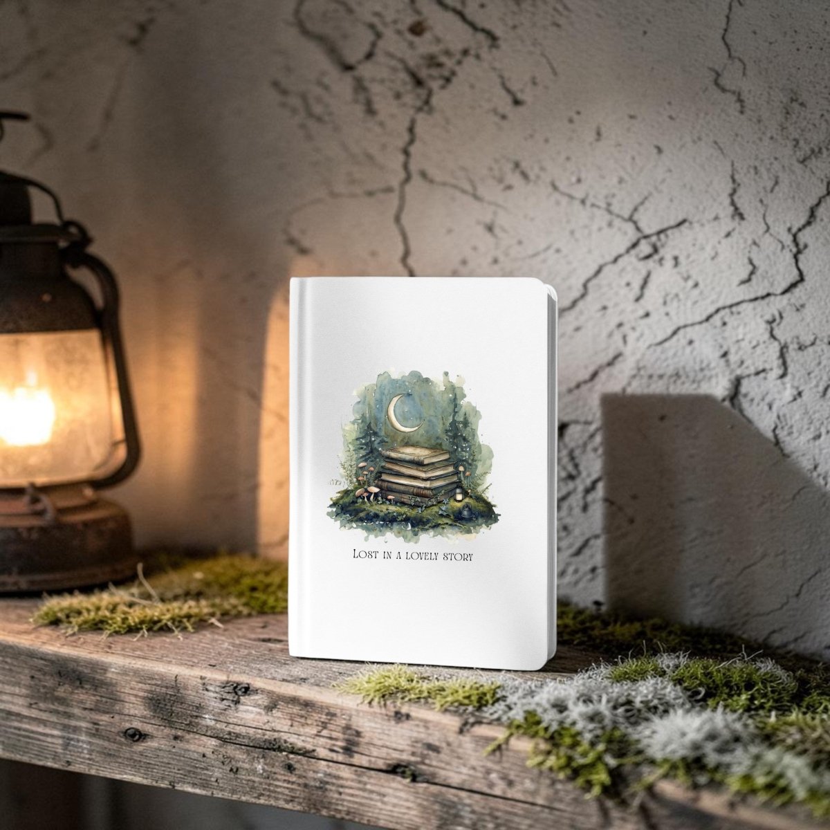 The Lost in a Lovely Story Journal by Mirabilia Boutique—a white hardcover featuring a moon and books illustration along with the words “LOVE IS A LONELY STORY”—stands on a rustic wooden shelf beside a glowing lantern, surrounded by moss and set against a textured stone wall, bringing woodland charm to any space.