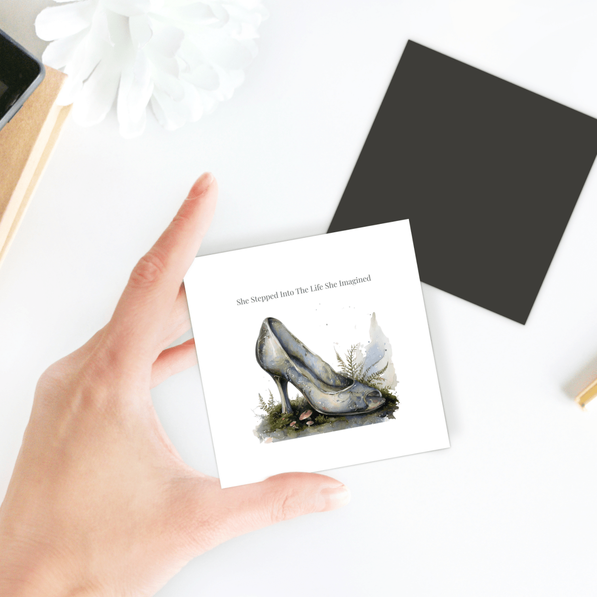 A hand holds the Mirabilia Boutique She Stepped Into the Life She Imagined Magnet, which features an illustration of a high-heeled shoe and the inspiring phrase “She Stepped Into The Life She Imagined!” Nearby, a black square, flower, and notebooks rest on a white surface, adding a whimsical fairytale flair to the decor.