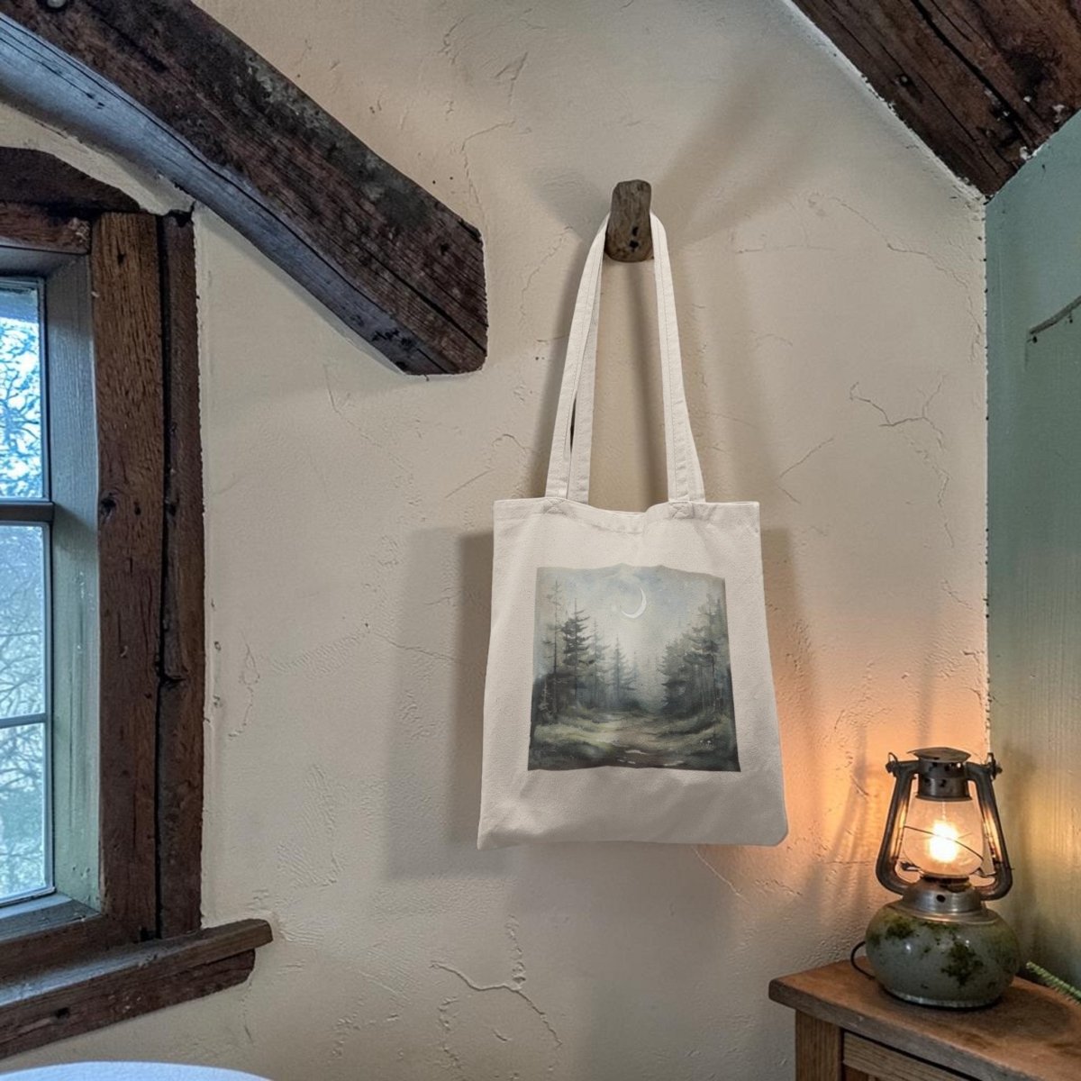 The "Some Paths Are Meant to Be Wandered" Tote Bag by Mirabilia Boutique, featuring a charming forest scene, hangs on a wooden peg beside a glowing lantern atop a small table in a rustic room with exposed beams and textured walls.