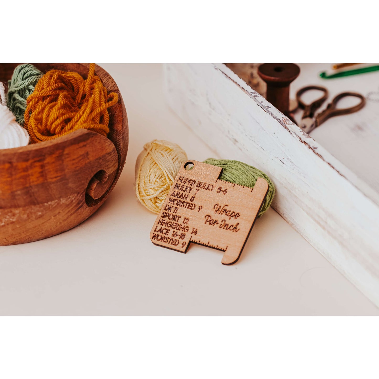 A wooden bowl holds mustard yellow, olive green, and white yarn. Linked to the gauge tool are Mirabilia Boutique's Knitter’s Pocket Tool Set—4 mini tools ideal for gifting and project bags. A white tray with scissors and a spool of thread is nearby, highlighting yarn gauge details.
