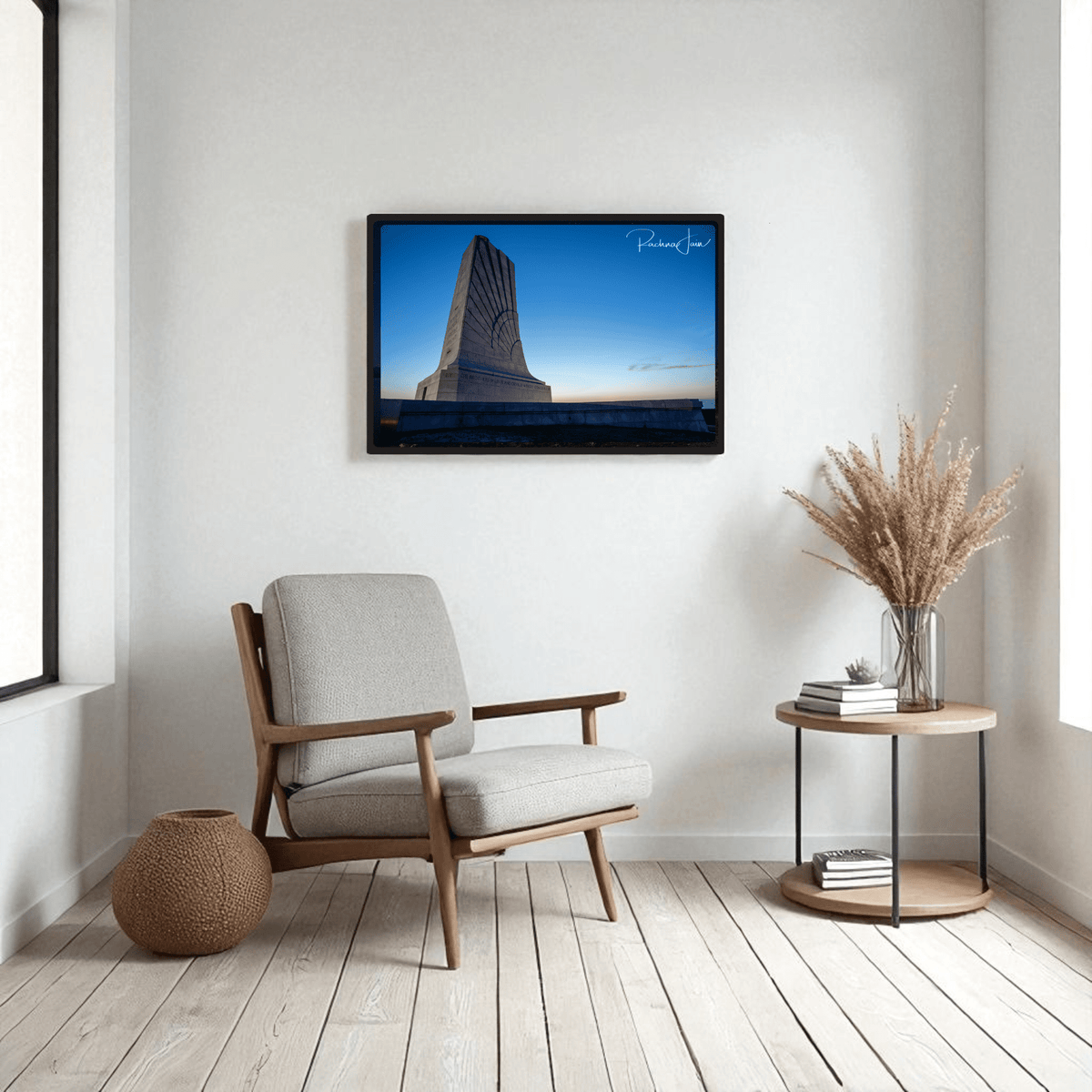 A modern living room with a cushioned chair, woven pouf, and round side table holding a vase of dried grass. On the wall hangs Mirabilia Boutique’s First in Flight Wright Brothers Monument Print. Light wood floors and white walls complete the look.