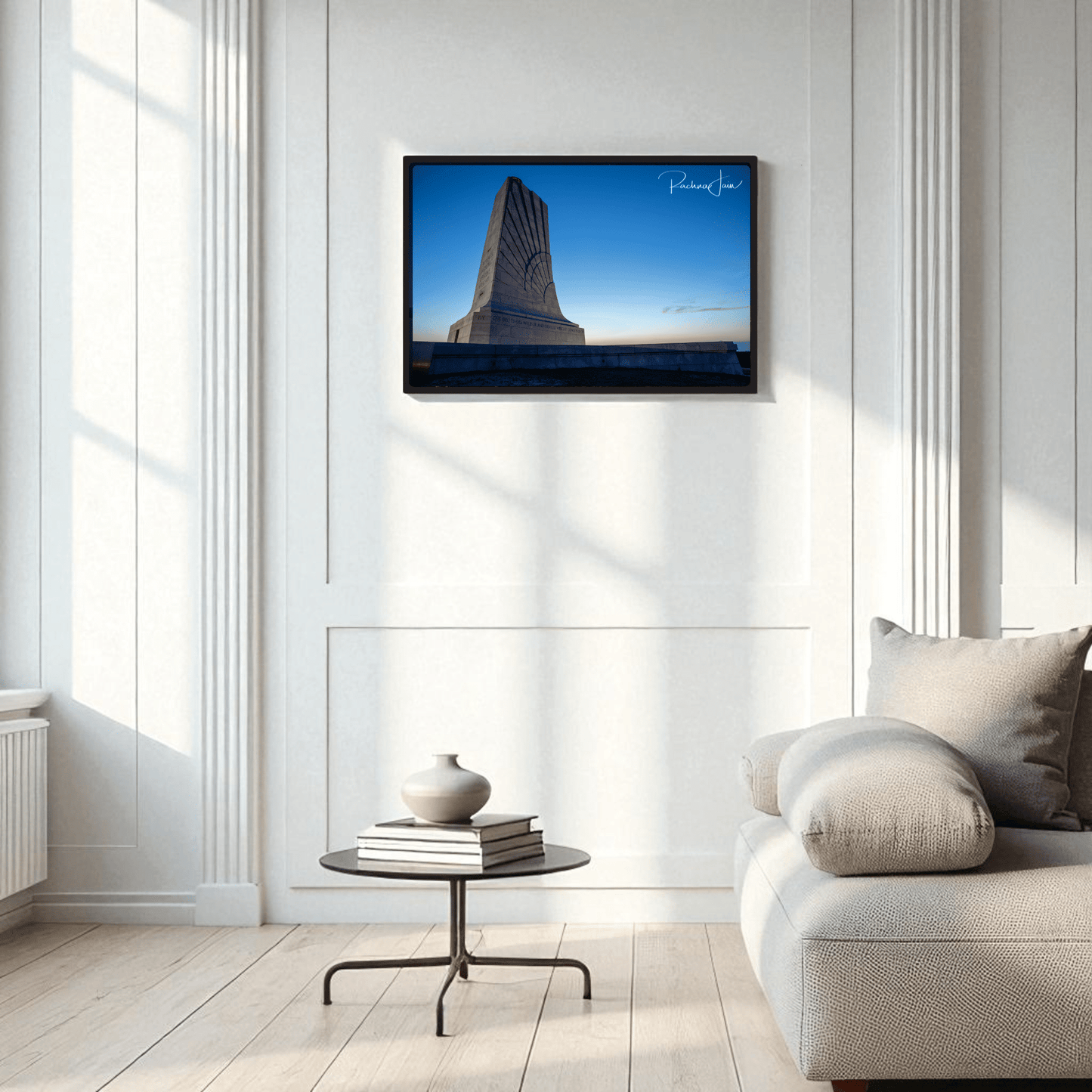 A modern minimalist living room with white walls and a beige sofa. A round coffee table with a vase sits in front, while Mirabilia Boutique’s Wright Brothers Monument Print | First in Flight Wall Art adds striking framed decor above the sofa.