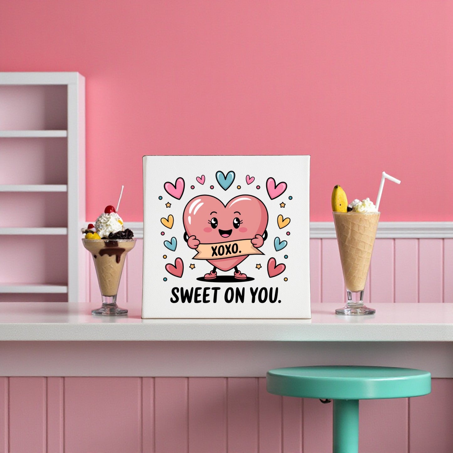 The Sweet On You XOXO Valentine Canvas by Mirabilia Boutique features a cute pink heart cartoon with a "XOXO" banner, surrounded by colorful hearts—perfect for Valentine’s decor or nursery art. Displayed in a retro pink dessert setting.