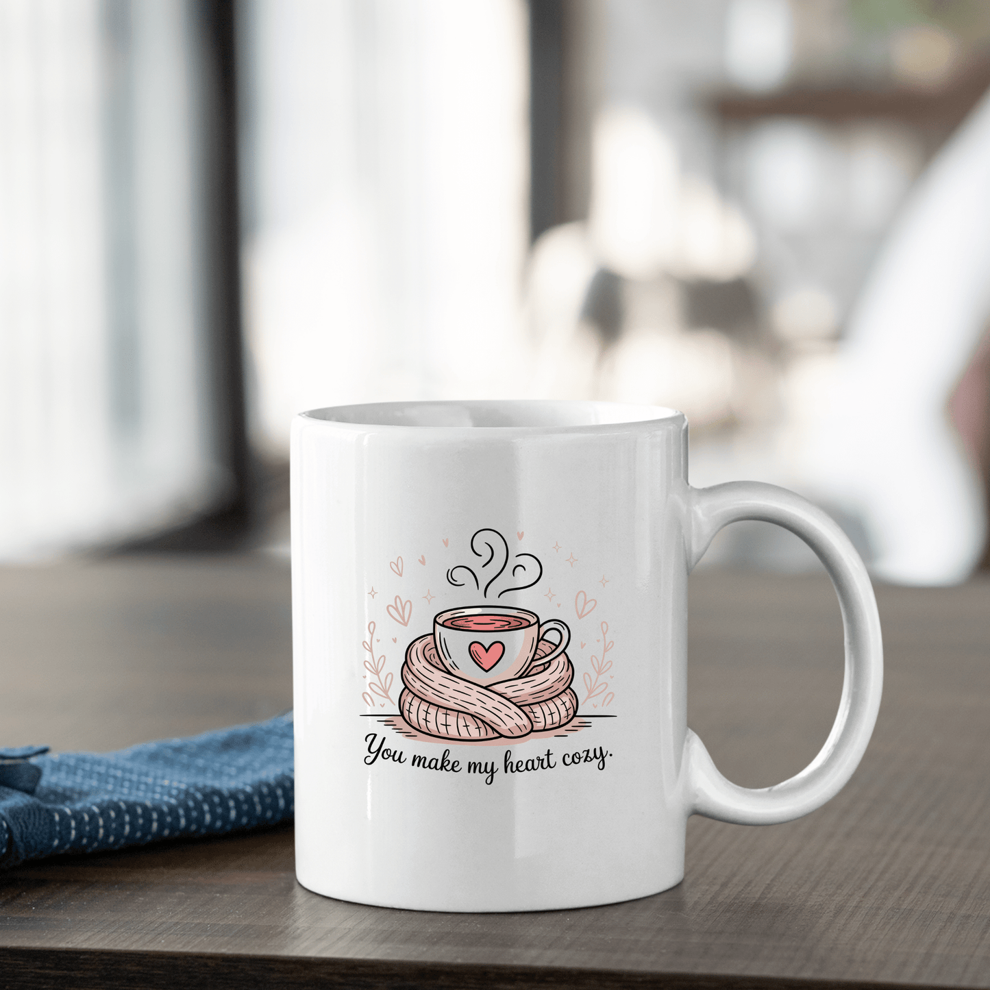 The You Make My Heart Cozy Mug by Mirabilia Boutique sits on a wooden table, featuring a steaming cup wrapped in a scarf with a heart and the text "You make my heart cozy," making it an ideal Valentine’s Day gift for coffee lovers.