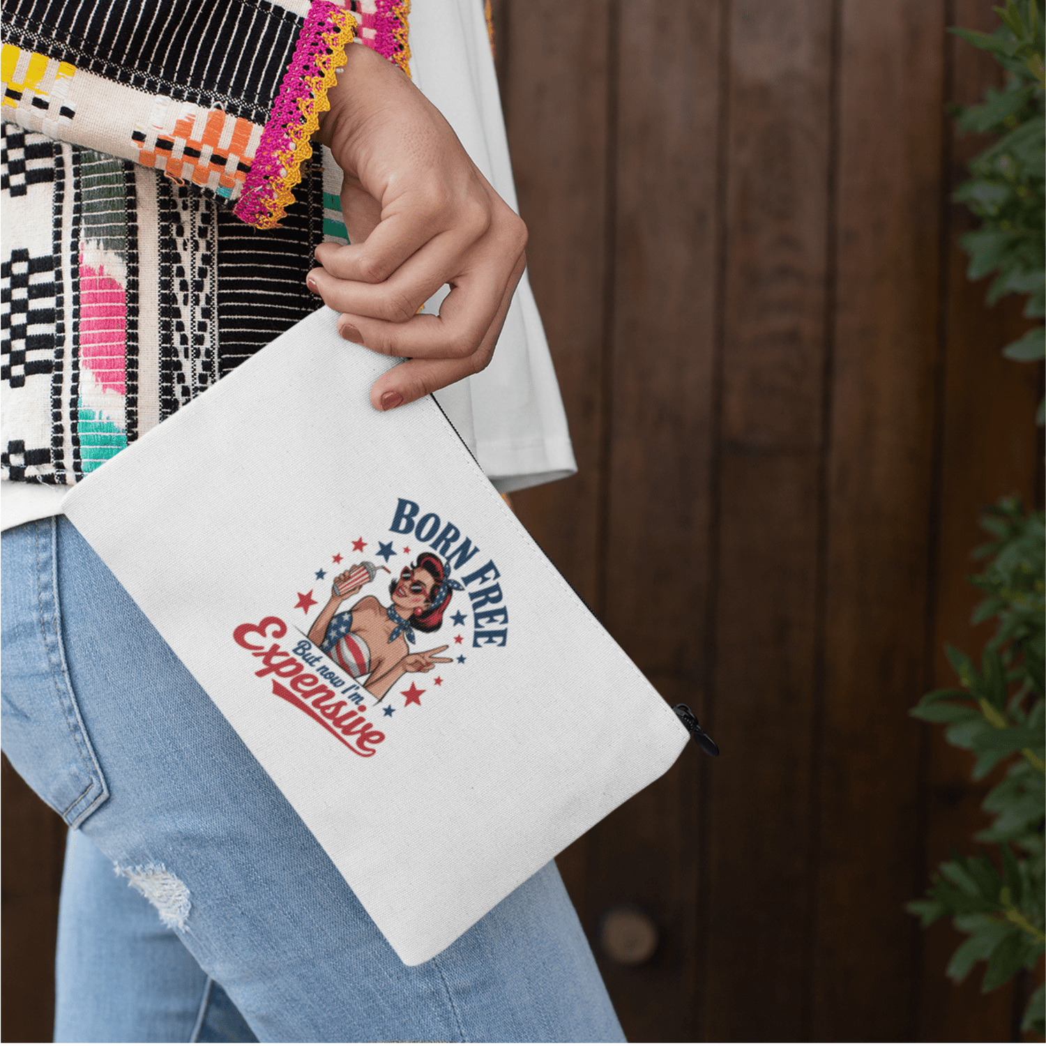 A white zippered pouch features a retro-style woman with sunglasses and a headband, surrounded by stars. Text reads: “Born Free but now I’m Expensive.”.