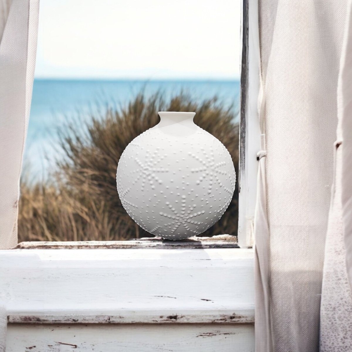 A round white vase with raised star patterns sits on a windowsill, framed by light curtains, with tall grass and the sea visible in the background.