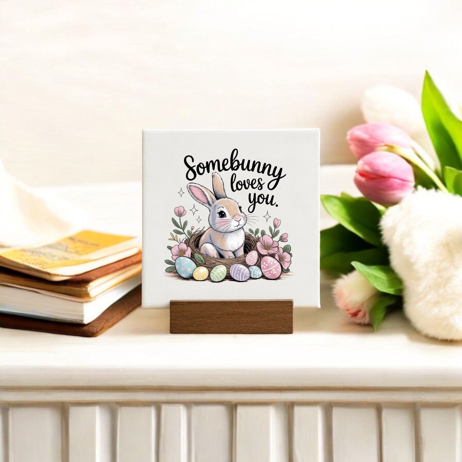 A small sign with a bunny, pastel eggs, and flowers reads "Somebunny loves you." It sits on a wooden stand next to stacked books and a pink tulip bouquet on a white surface.