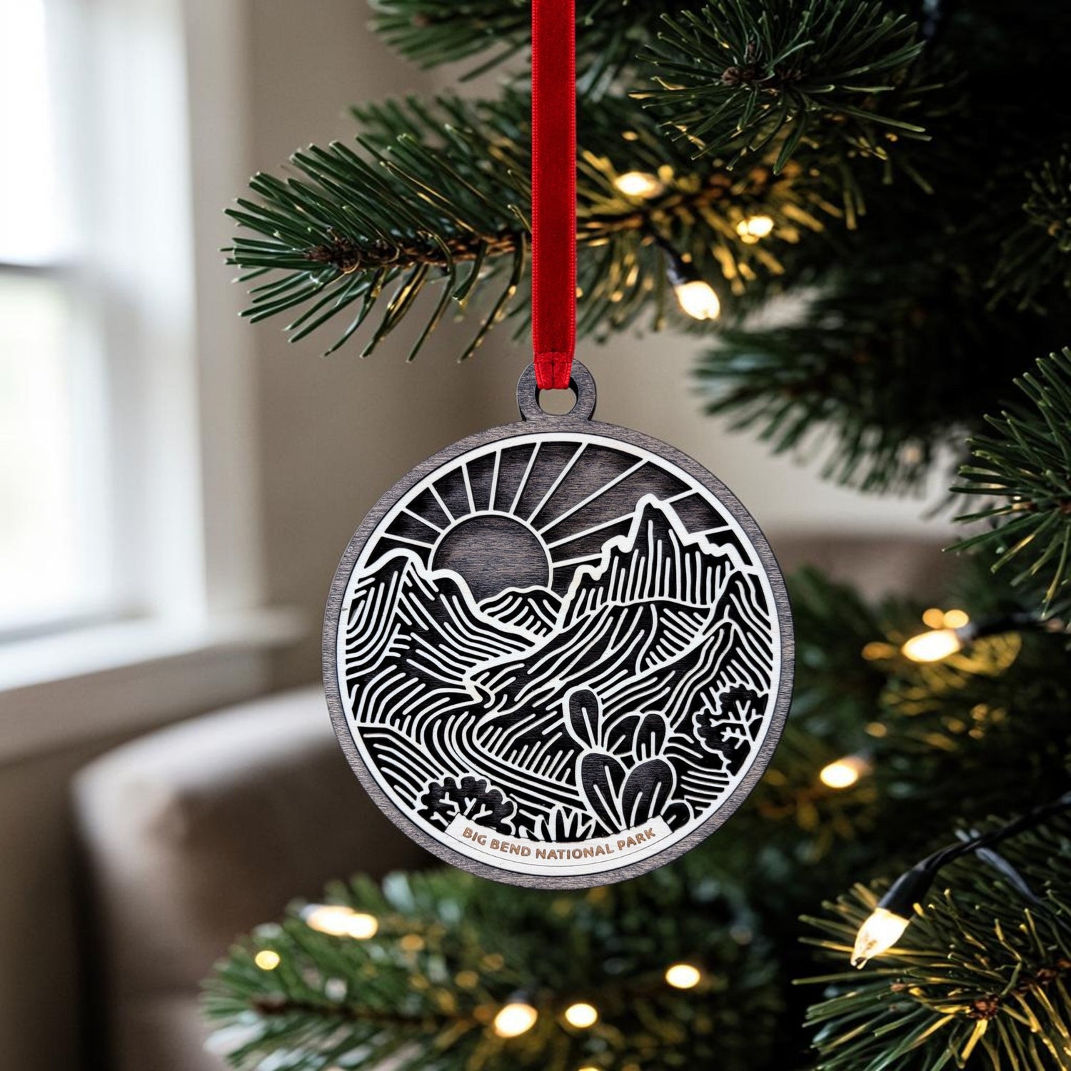 A round, wooden ornament with a laser-cut design of mountains, trees, and the words "Grand Teton National Park" hangs on a string outdoors, with snow-covered trees blurred in the background.
