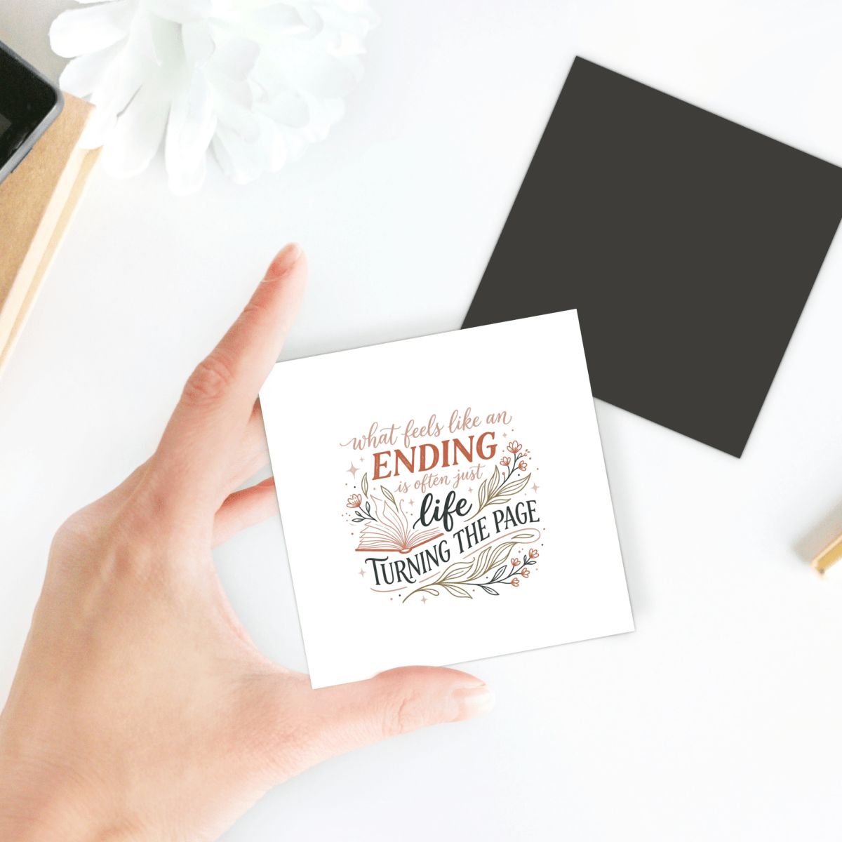 A hand holds a card with the quote, “What feels like an ending is often just life turning the page,” with floral decorations. A black square, plant, and notebook are on a white desk in the background.