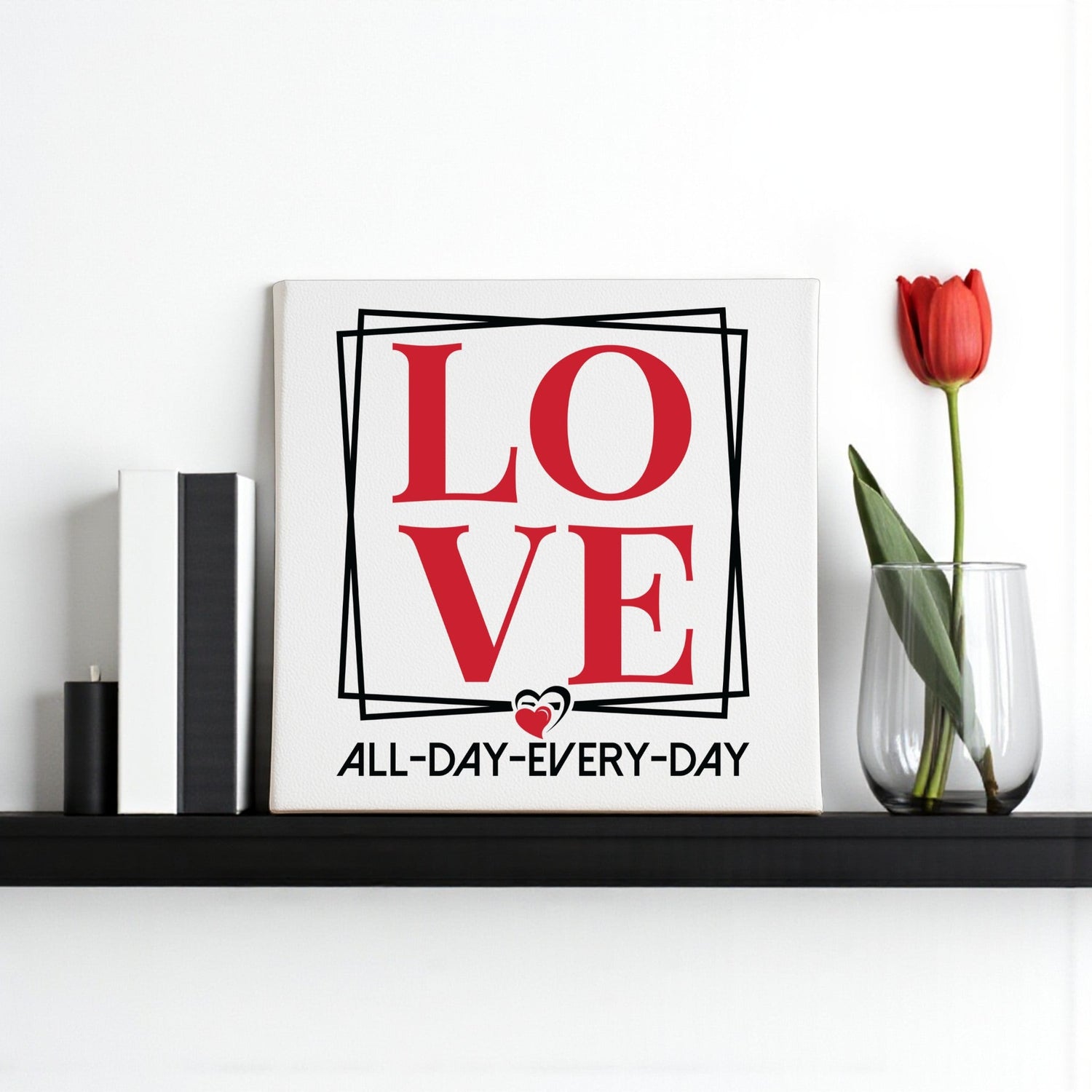 A shelf with a framed print that says “LOVE ALL-DAY-EVERY-DAY” in red and black, next to two books and a single red tulip in a clear glass vase.
