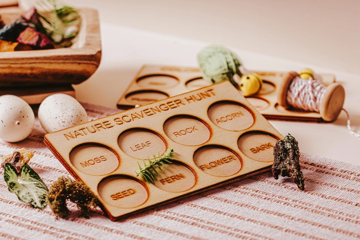 A wooden Nature Scavenger Hunt board with labeled sections for moss, leaf, rock, acorn, seed, fern, flower, and bark, surrounded by nature items and craft supplies on a striped cloth.
