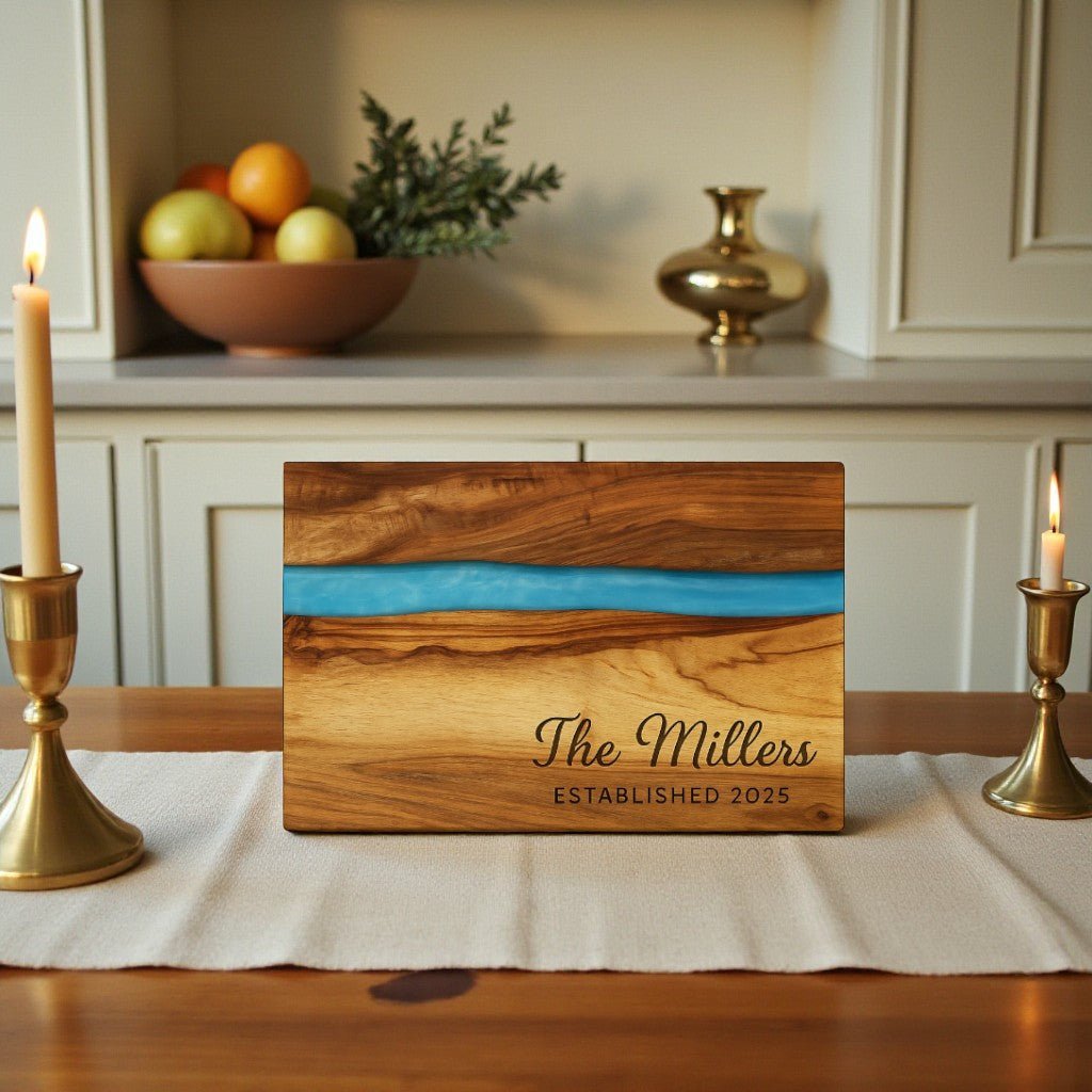 A personalized wooden sign with a blue resin inlay reads "The Millers ESTABLISHED 2025." It sits on a table between two lit candles, with fruit and a vase in the background.