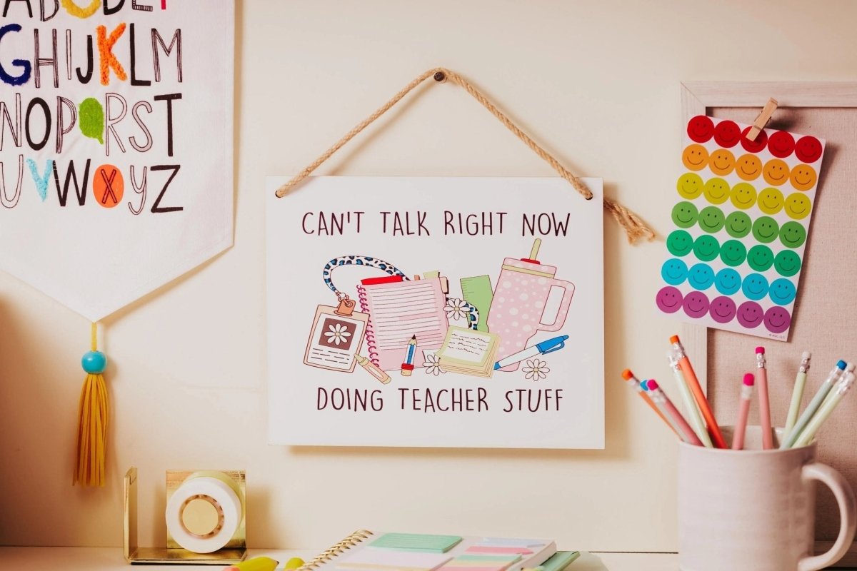 A classroom desk with colorful pens, a smiley-face pop-it, and a sign that reads, "Can't talk right now, doing teacher stuff," surrounded by notebooks and supplies.