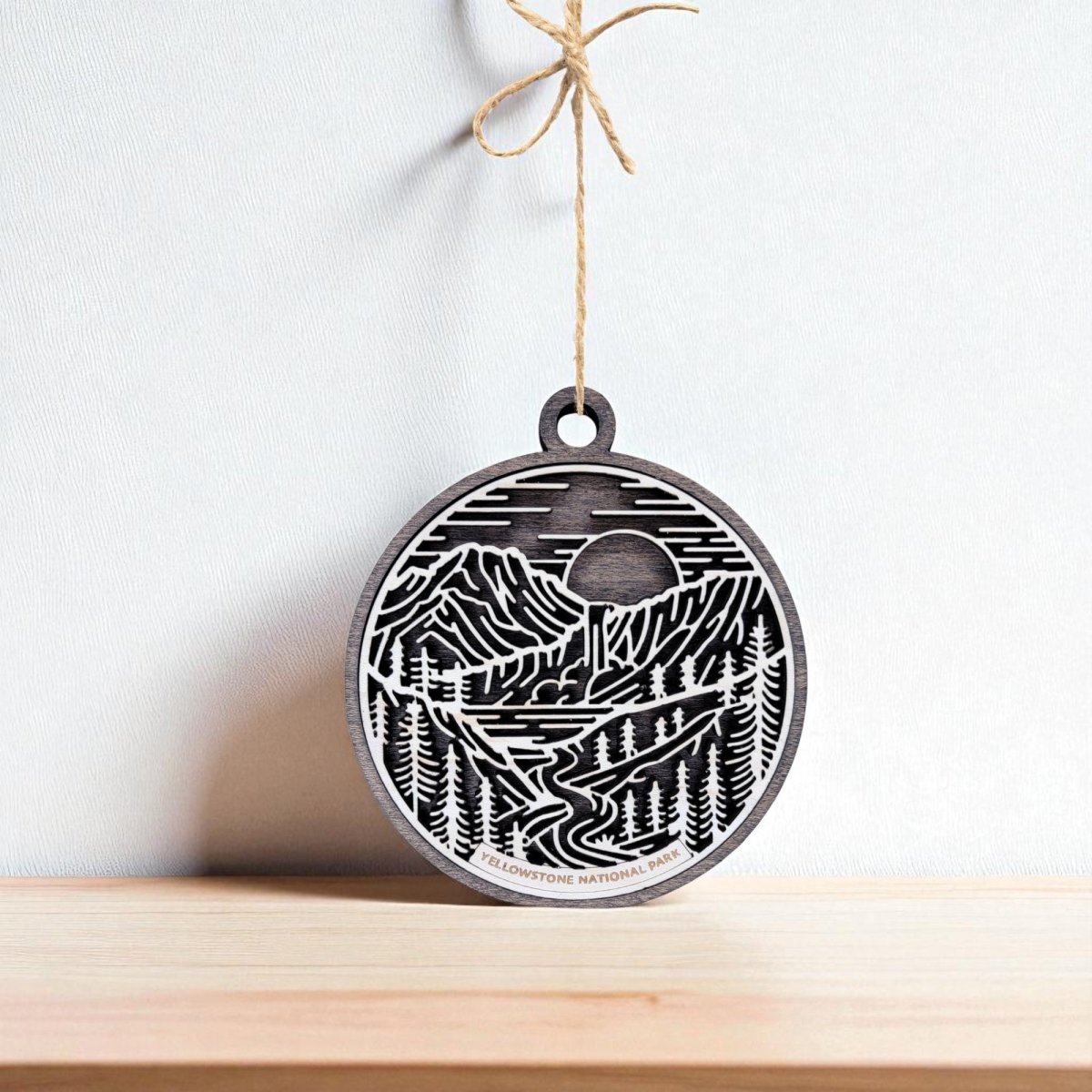 A round wooden ornament with a laser-cut landscape design of mountains, trees, and a river, hanging by a twine string against a white background above a light wooden surface.