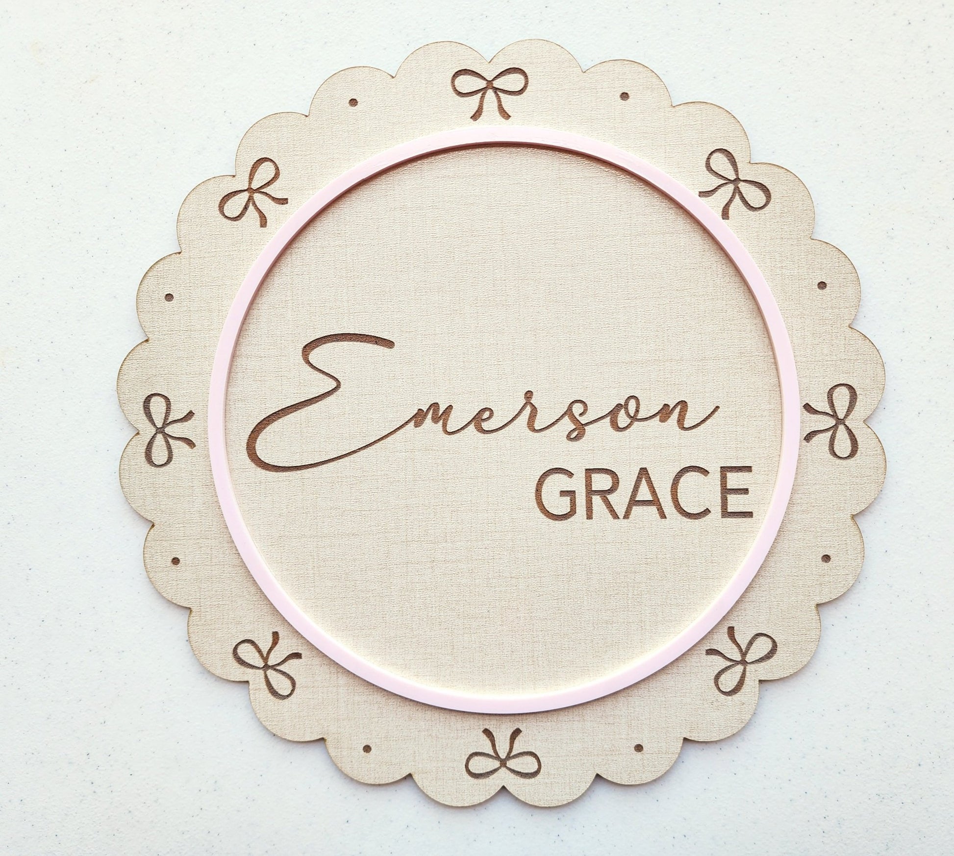 The Mirabilia Boutique Personalized Baby Name Shelf Sign features scalloped edges, delicate bows, custom name in script and bold text, and a thin pink border—a charming beige wooden nursery decor or newborn gift.